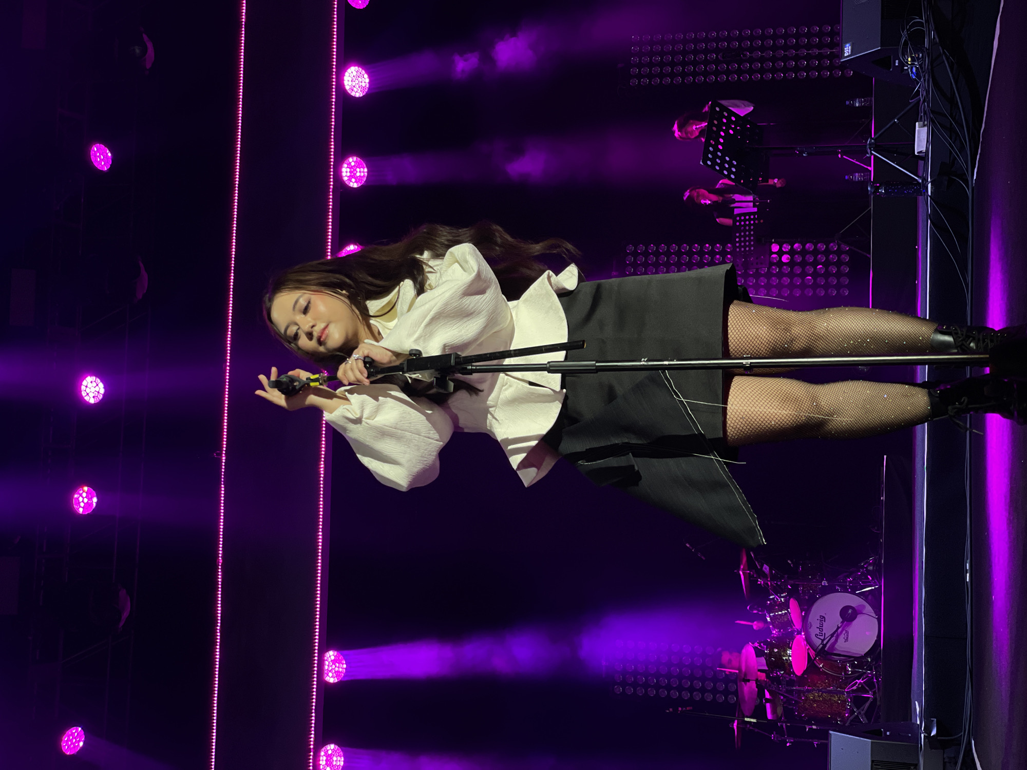 A woman is singing on stage in a concert venue in Salembaran, Indonesia. She has long, dark hair and is wearing a white blouse with voluminous sleeves and a dark skirt. She is holding a microphone in her left hand and her right hand is raised. She is wearing sparkly fishnet stockings. The stage is lit with purple lights and spotlights. A drum set is visible in the background, along with stage equipment and a music stand with two figures behind it.