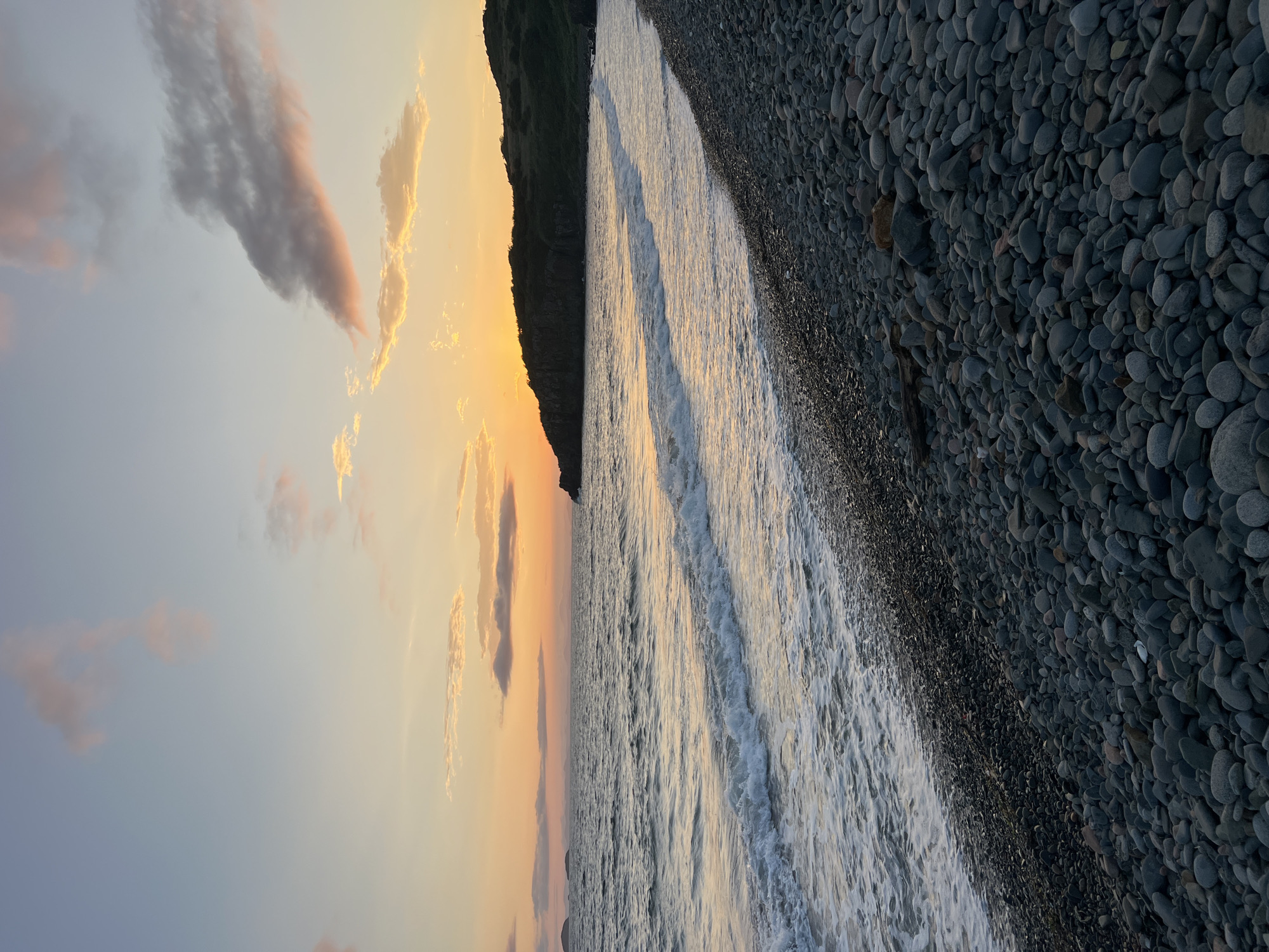 The image depicts a pebble beach meeting the ocean at sunset. The sky transitions from a pale blue at the top to a warm orange hue near the horizon, with scattered clouds tinged pink and grey. Gentle waves with white foam break along the shore. The beach is covered in smooth, dark grey pebbles of various sizes, with some larger rocks and a piece of driftwood visible in the foreground. In the distance, a dark, hilly coastline forms a silhouette against the sunset. The location is identified as Reineke, Russia. There are no people or animals present in the image.
