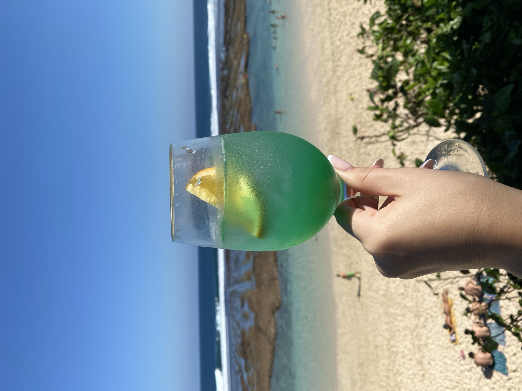 A hand holds a glass of green liquid with ice and an orange slice in front of a beach scene in Kuta Selatan, Indonesia. The beach has light-colored sand and is dotted with several people sunbathing or wading in the shallow, turquoise water. Beyond the water, a darker blue ocean stretches to the horizon under a clear blue sky. Rocky formations are visible in the shallow water near the shore.