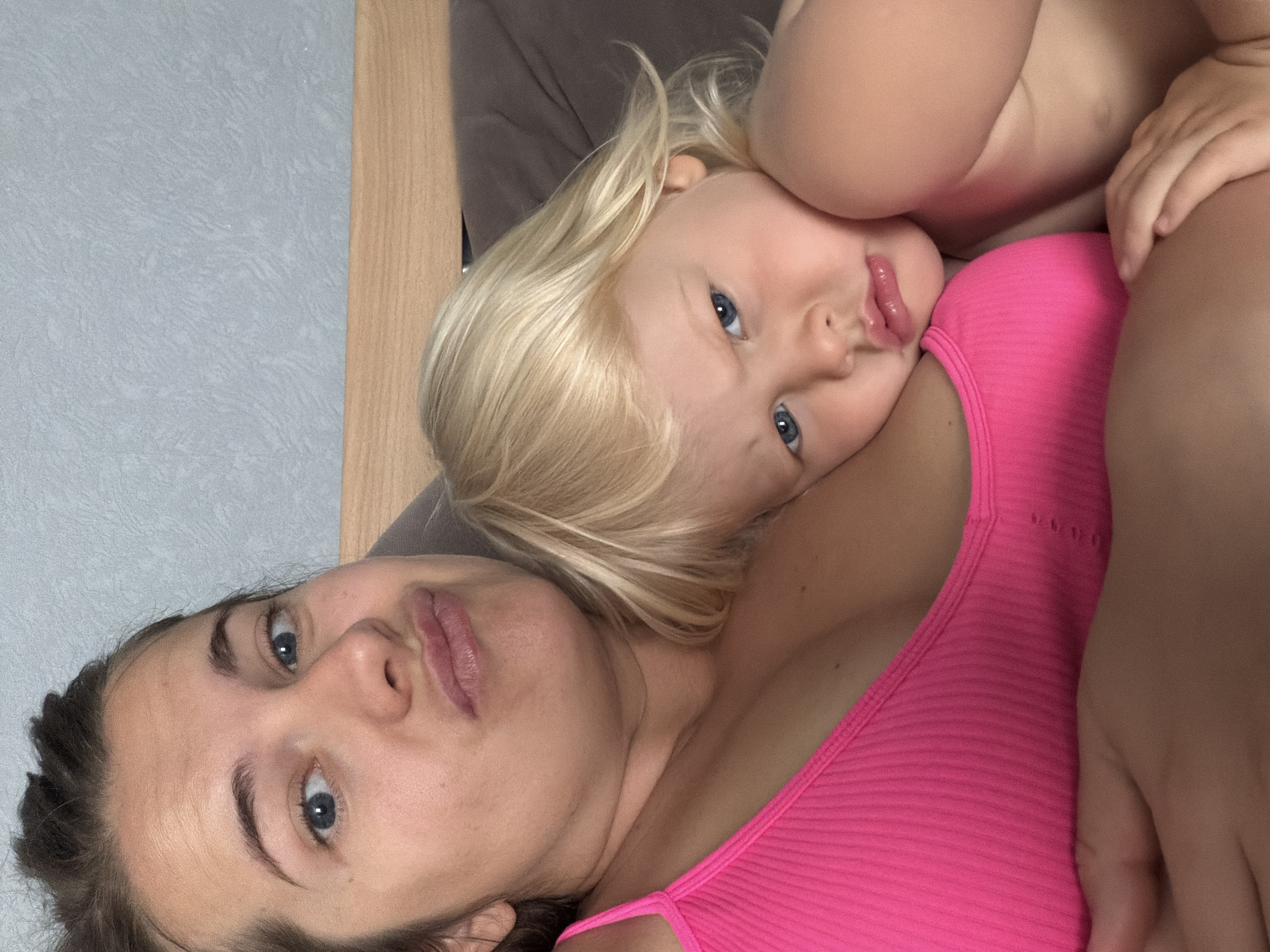 A woman with blue eyes and dark hair pulled up is making a pouting face at the camera. A young child with blonde hair and blue eyes is resting their head on the woman's chest, looking towards the camera with a neutral expression. They are both in a close-up shot. The woman is wearing a bright pink ribbed tank top. The background shows a textured light blue wall and a wooden headboard. The scene appears to be indoors, likely in a bedroom. There is no indication of the location being Sochi, Russia.