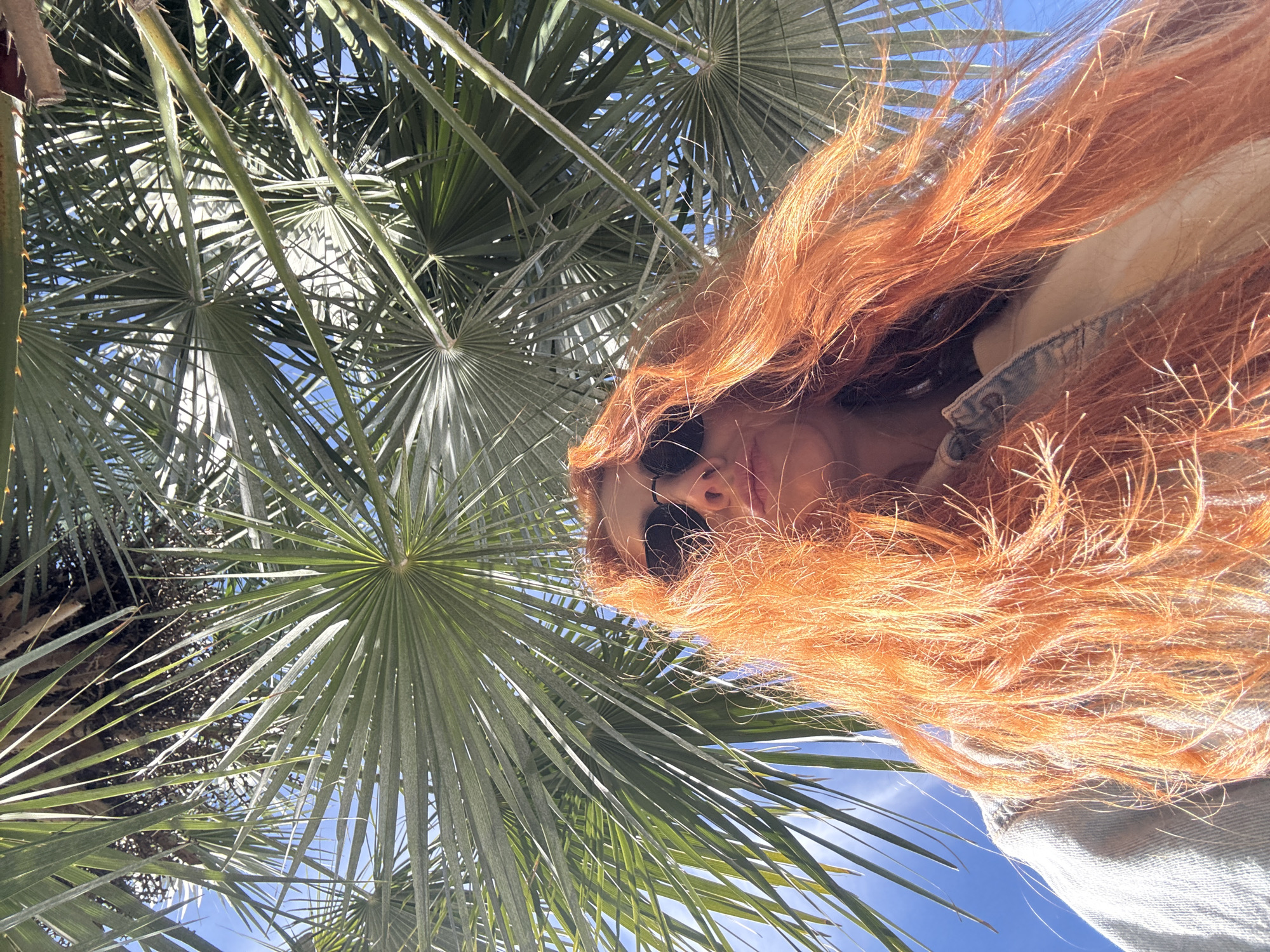 The photograph presents a low-angle view looking up into a fan palm and the face of a person with long, red-orange hair. The scene is sunlit. The person is wearing sunglasses, and part of a light-colored jacket with a denim-looking texture is visible. The sky is blue, visible through the palm fronds. The context clues from the location prompt suggests that this scene is located in Budva, Montenegro, an urban location. The interaction suggested is the person photographing themself or being photographed from below.