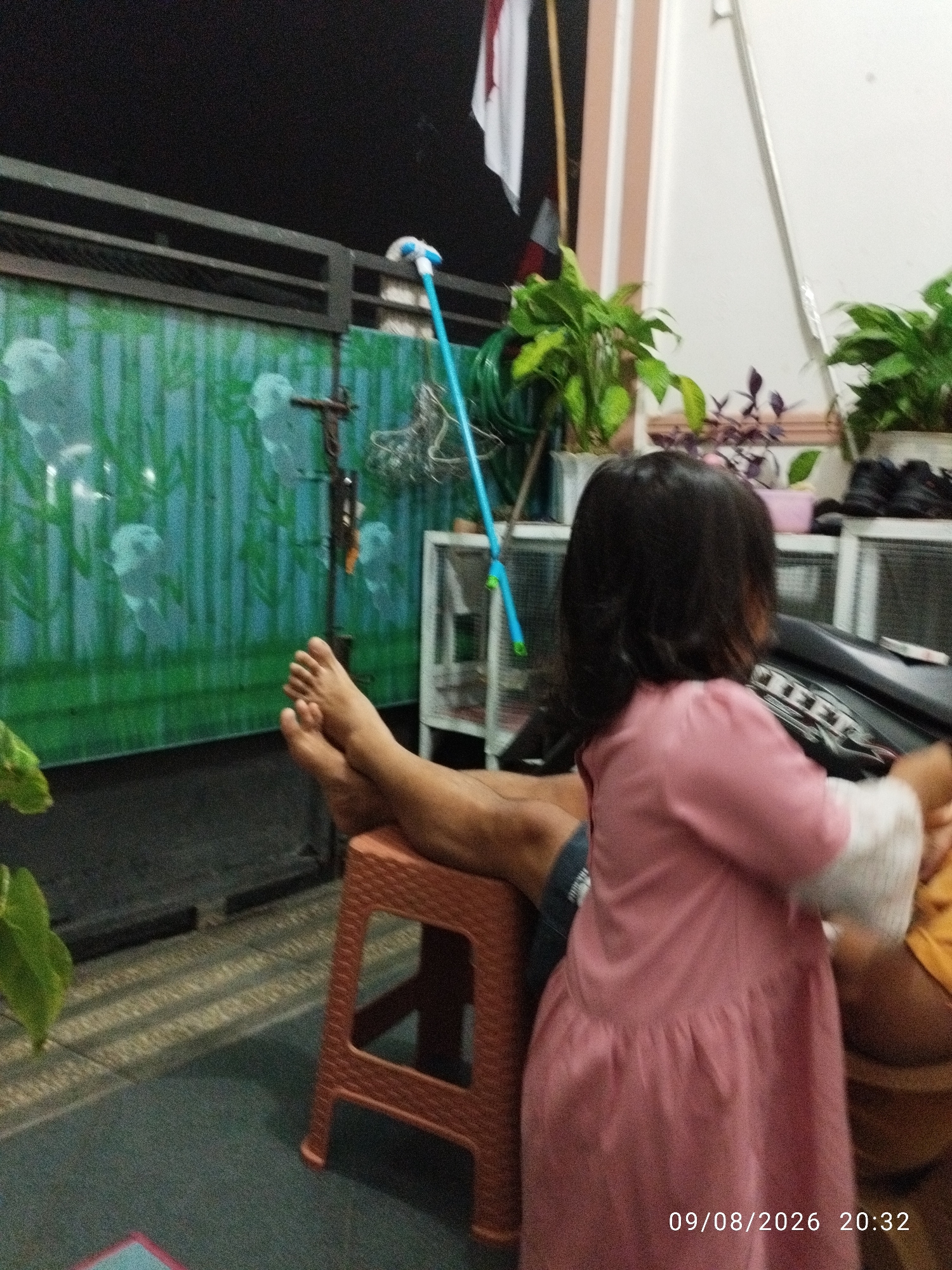 A person's bare feet are propped up on a reddish-brown plastic stool, positioned on a dark tiled floor. A child in a pink dress is standing in front of the person, facing away from the camera. The background features a green wall with a bamboo-like pattern, a dark metal fence, and various potted plants. A blue mop is leaning against the wall. The timestamp 09/08/2026 20:32 is visible in the lower right corner. The setting appears to be an outdoor residential area, possibly a yard or patio, in Bandar Lampung, Indonesia.