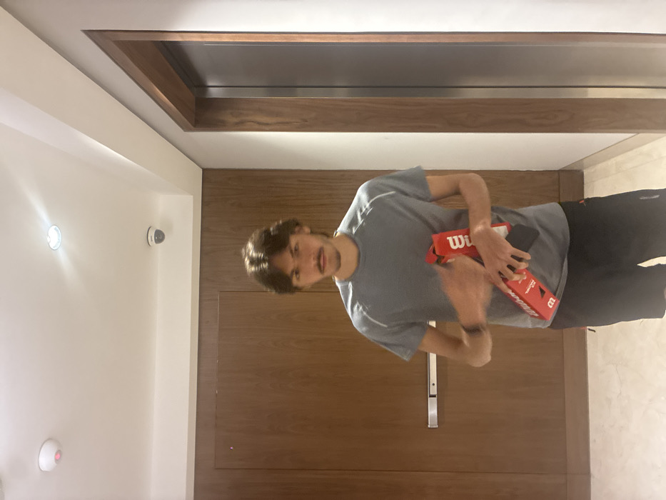 The image depicts a male subject standing in an indoor hallway of a building in Mexico City, Mexico. The subject, appearing to be a young adult with dark, medium-length hair and a mustache, is positioned centrally, facing directly forward. He is wearing a gray short-sleeved athletic shirt with white trim and black athletic shorts. A dark wristband is visible on his left wrist. The subject holds a red rectangular container of Wilson brand tennis balls; his left hand is blurred, indicating motion, while his right hand firmly grasps the container.
The background features a light-colored floor and a white ceiling with two recessed light fixtures, a red circular smoke detector, and a white dome-shaped security camera. To the subject's left is a large, dark brown wooden door equipped with a horizontal silver push bar. To the subject's right is a dark metallic door, likely an elevator, framed by dark brown wood trim set against a light-colored wall. The scene is uniformly lit by artificial light.