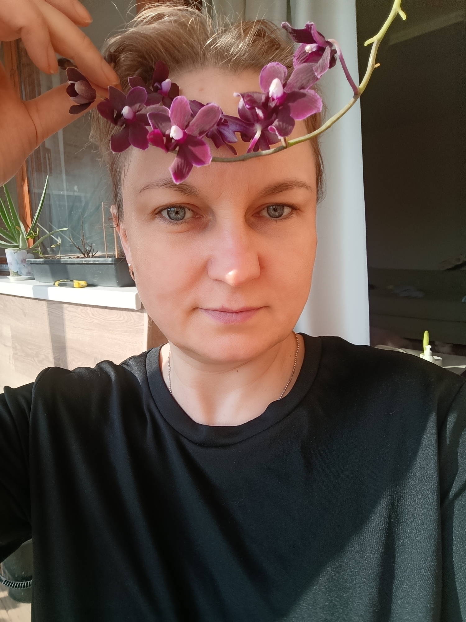 A woman is holding a stem of purple orchids in front of her face, just above her forehead. She has short, light-colored hair and is wearing a black t-shirt and a silver necklace. The background includes a window with plants on the windowsill, and a doorway in the back. The location is Марковский, Russia.