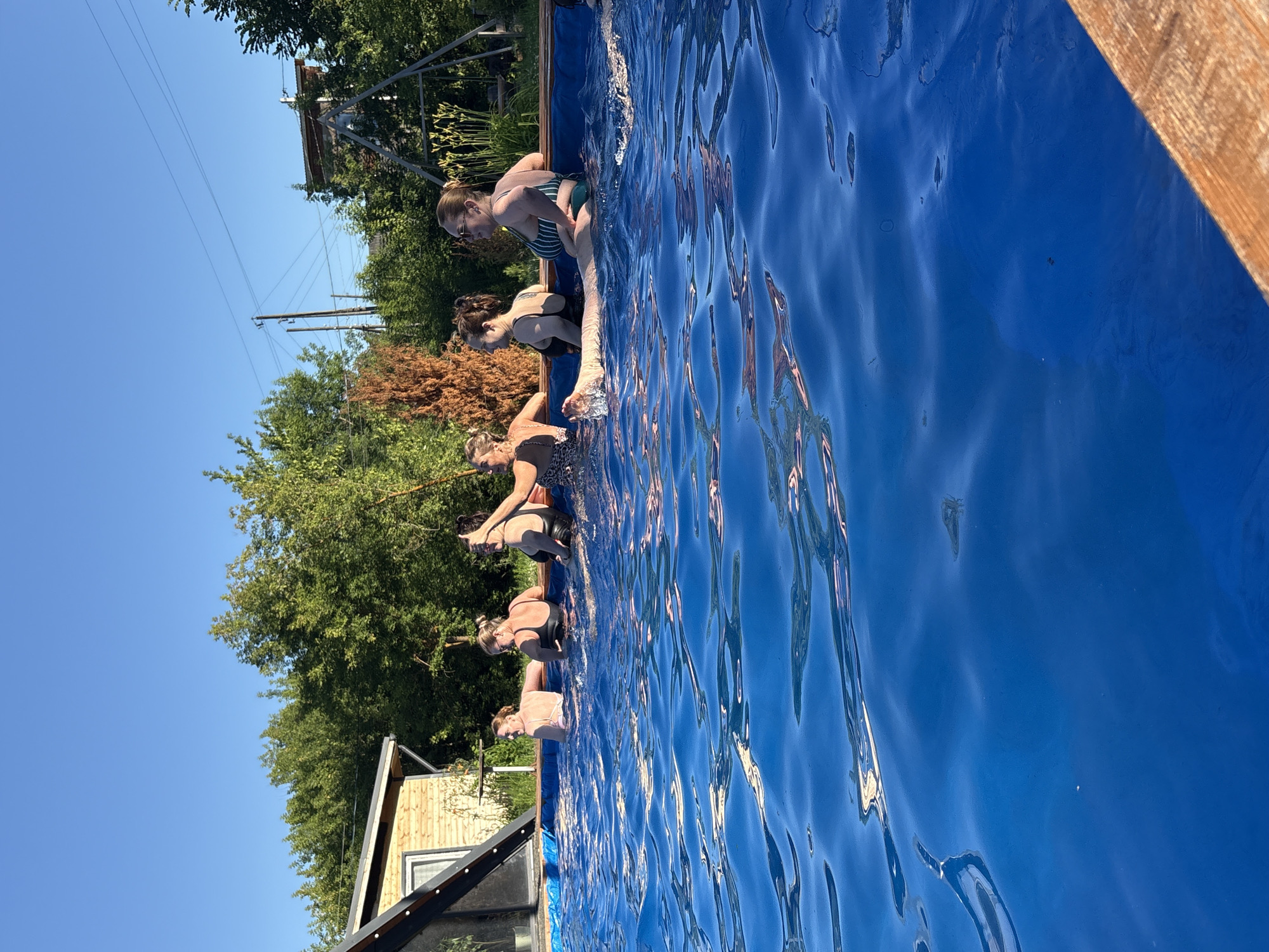 Six individuals are partially submerged in a blue swimming pool on a sunny day. The pool is set within a yard with a wooden fence or deck visible in the foreground. In the background, there is a wooden building with a slanted roof, green trees, and power lines. The individuals are dressed in swimwear, and some appear to be interacting with each other. The scene suggests a recreational activity taking place outdoors.