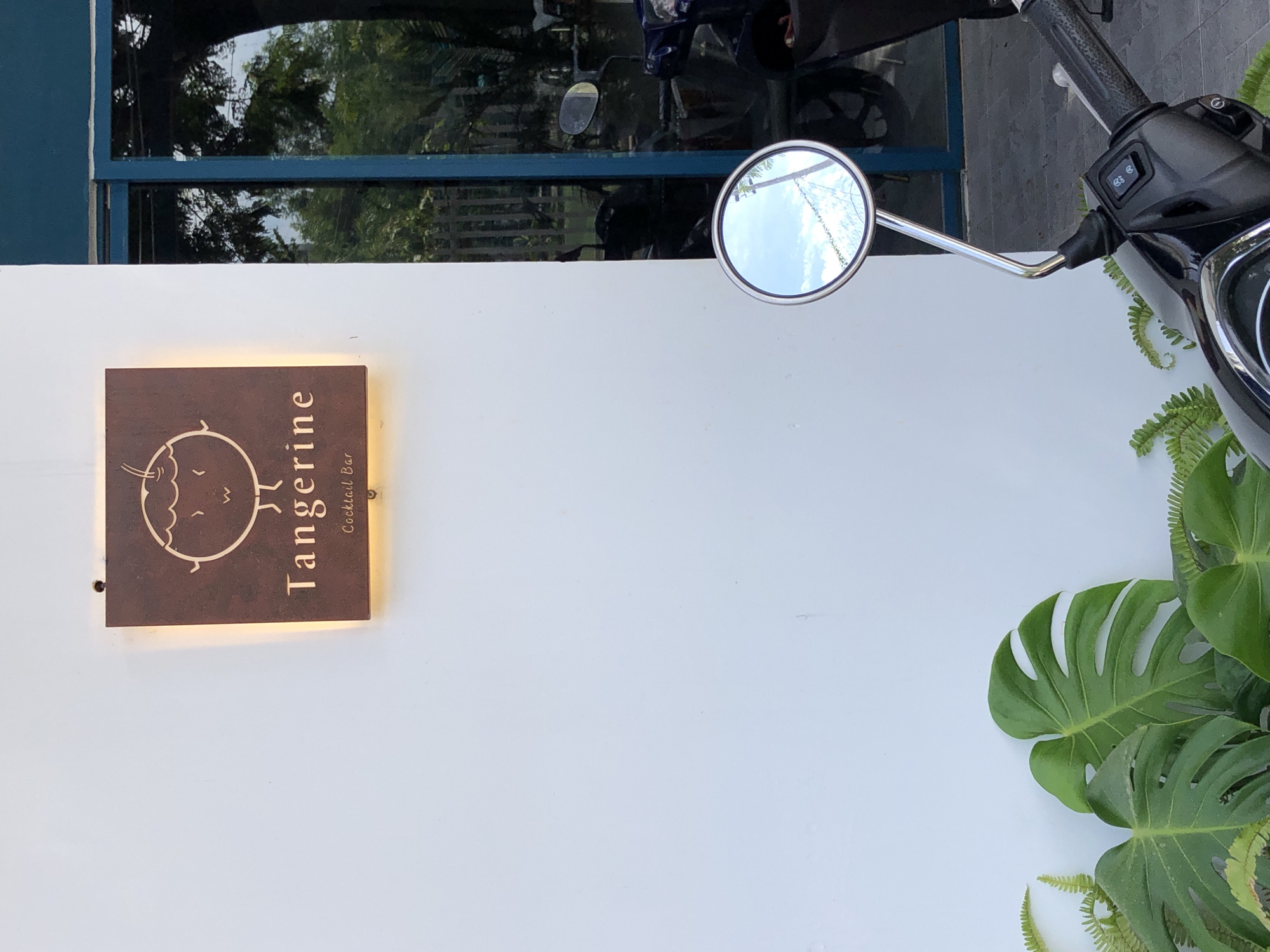 An outdoor shot captures a white wall with a sign for "Tangerine Cocktail Bar" featuring a stylized orange character. To the right, a blue-framed window reflects lush green trees. In the foreground, a motorcycle's handlebars and a circular mirror are visible, reflecting a bright blue sky with scattered clouds. Several large, green leaves, likely from a Monstera plant, and some ferns are arranged at the base of the white wall. The scene suggests a modern establishment in a city, possibly in Đà Nẵng, Vietnam, given the presence of tropical foliage and common urban elements like motorcycles.