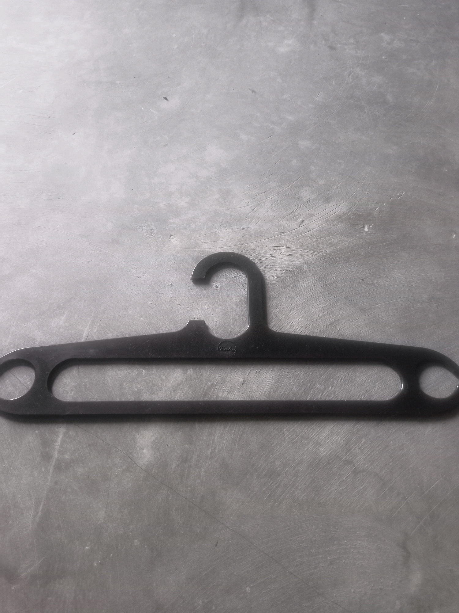 A black plastic clothes hanger lies horizontally on a textured, gray surface, likely a table or countertop. The hanger is centrally located in the frame and has a standard shape with a hook at the top and extended arms with circular openings at the ends. There are faint markings on the hanger, possibly a brand name, that are not clearly legible. The surface beneath the hanger displays a matte finish with visible scuff marks and subtle variations in tone, suggesting it is made of a material like metal or concrete. The lighting in the scene is neutral, casting soft shadows that highlight the texture of the surface and the form of the hanger. No people or any other objects are visible in the image. The scene is devoid of any indication of its specific location within Karangtalun, Indonesia.