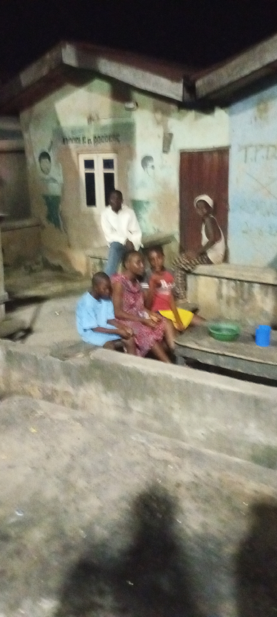The image captures a nighttime scene in Adankolo, Nigeria, focusing on a group of people gathered outside a building. The building appears to be a house or community center with a light-colored facade adorned with murals of people. A window with white panes is visible in the center of the facade.
In the foreground, several individuals are seated on a concrete ledge. A young boy in a light blue shirt is on the far left, looking down. Next to him, a woman in a patterned dress is seated. To her right, a young girl in a red shirt and yellow skirt is also seated. Further to the right, a woman wearing a white headscarf and a white tank top sits on the ledge, looking towards the camera.
Standing behind the seated group, near a dark wooden door, is a man in a white long-sleeved shirt and blue trousers. The ground in front of the building is rough and uneven. To the right of the seated individuals, a green bowl and a blue plastic cup are placed on the ledge. The overall lighting suggests it is evening or night, with artificial illumination casting shadows. The background shows more of the building's exterior, including what appears to be a door on the right side and some indistinct writing or art on the wall.