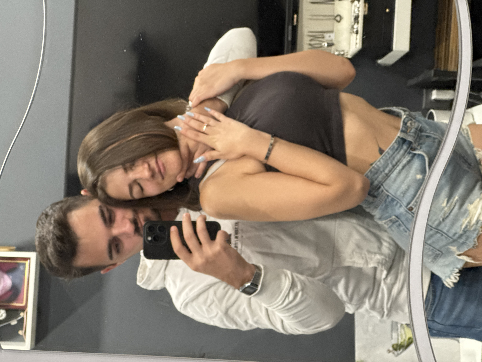 A couple takes a mirror selfie in what appears to be a home or dressing room. The man, wearing a white hoodie and a watch, holds a smartphone to capture the image. The woman, dressed in a dark crop top and denim shorts, has her eyes closed and a slight smile as the man places his hand on her neck. Both individuals have jewelry on their hands, with the woman wearing a ring and bracelet. The background shows a mirror reflecting the room, which includes a framed picture on a shelf, a dark wall, and a display case filled with jewelry. The lighting is dim, and the overall atmosphere is intimate.