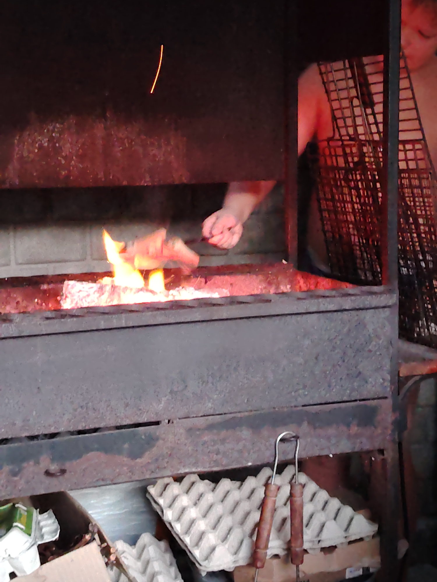 A person is grilling food over a fire inside a barbecue pit. The scene is indoors or under a cover, with a brick wall behind the pit. To the right of the pit, a metal grilling rack is visible, and a person's arm and shoulder are also present. In the foreground, the bottom of the barbecue pit contains hot coals and flames. Below the pit, there are cardboard egg cartons and other miscellaneous items. Two wooden-handled grilling tools are leaning against the egg cartons. The image is somewhat blurry and low-resolution, with a reddish tint from the fire and lighting. The location is unknown.