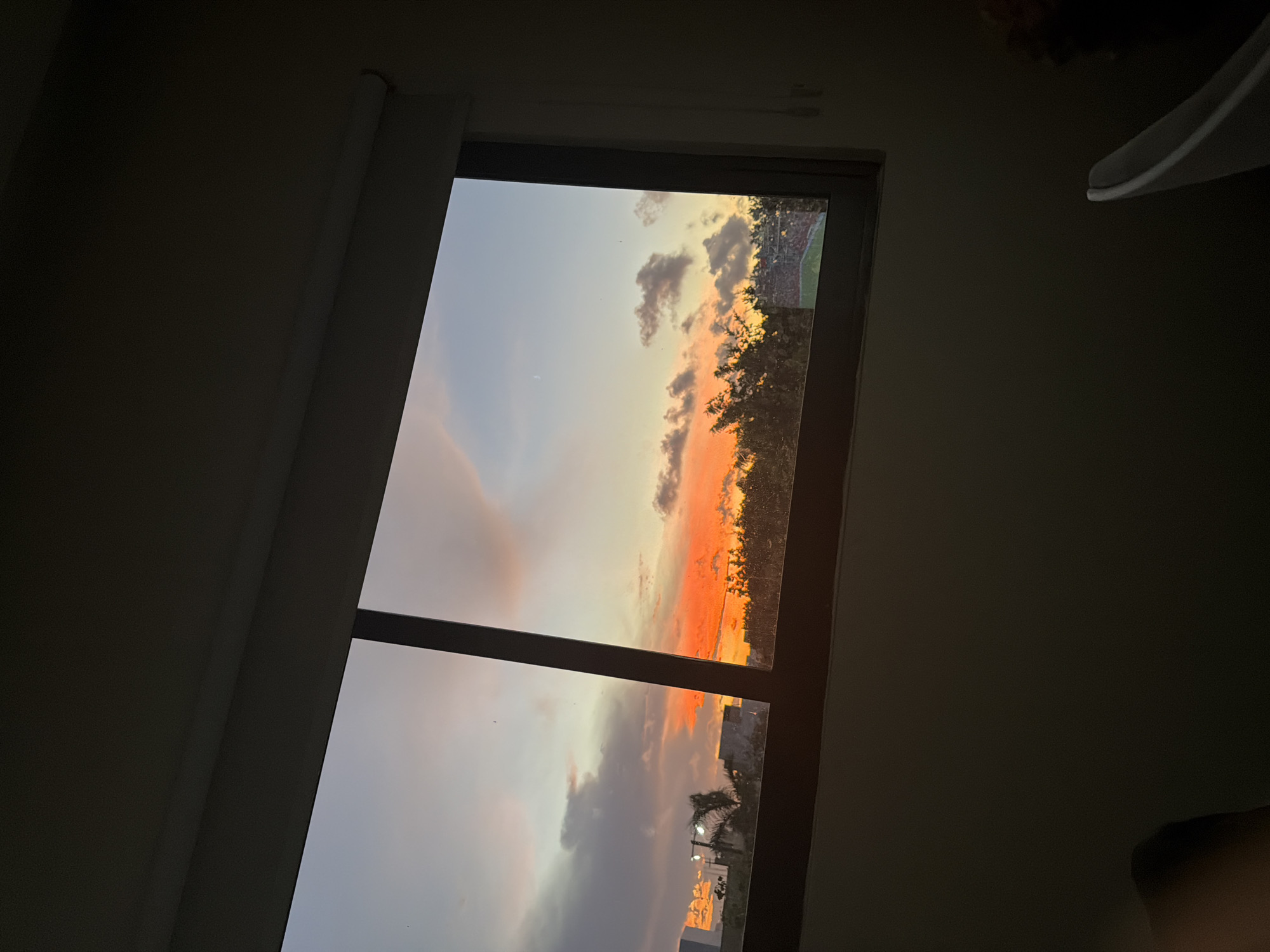 A late afternoon sky, illuminated by the setting sun, is visible through a large window divided by a dark vertical mullion. The sky displays a gradient of colors, from a pale blue at the top to vibrant oranges and yellows near the horizon, interspersed with scattered grey clouds. Below the sky, the silhouettes of buildings and lush green trees create a stark contrast against the bright sky. In the distance, a stadium with red seating is discernible through the trees. The foreground is predominantly dark, with the edge of a white chair visible on the right. The setting appears to be an interior space in Cancún, Mexico, given the tropical foliage and architectural hints.