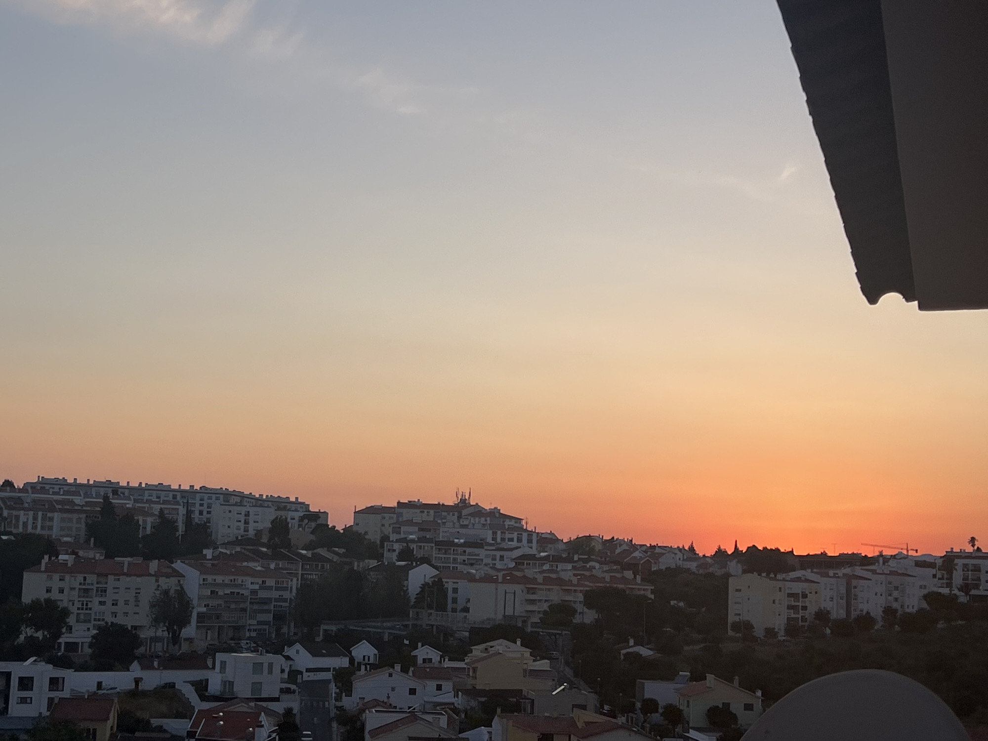 The image captures a panoramic view of the cityscape of Cascais, Portugal, during a sunset. The sky transitions from a pale blue at the top to vibrant shades of orange and red near the horizon, indicating the end of the day. Below the sky, a densely populated residential area is visible, characterized by numerous buildings of varying heights and architectural styles. Many buildings are multi-story apartments with white and beige facades, some featuring red-tiled roofs. The terrain appears hilly, with buildings situated on slopes, creating a tiered effect. Lush green trees and vegetation are interspersed among the buildings, particularly on the lower slopes. In the background, some larger structures and what appear to be communication towers can be seen on the highest points of the city. The foreground is partially obscured by a dark, angled structure, likely part of a balcony or rooftop, suggesting the photo was taken from an elevated vantage point. The overall scene depicts a peaceful, urban setting bathed in the warm light of the setting sun.