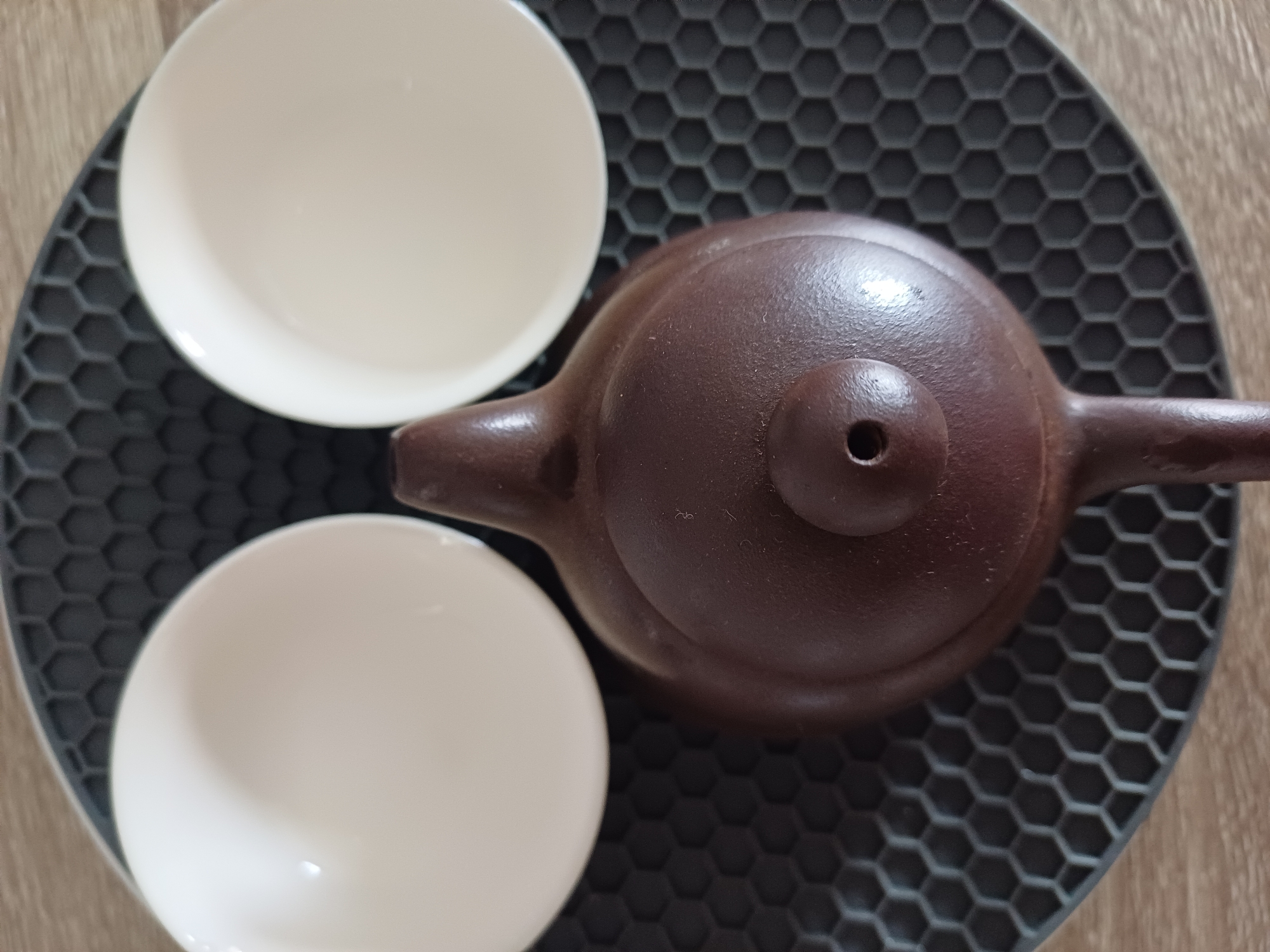 Two white teacups and a brown teapot sit on a gray, hexagonal patterned mat. The mat rests on a light brown surface. The photograph was likely taken in Gošići, Montenegro.