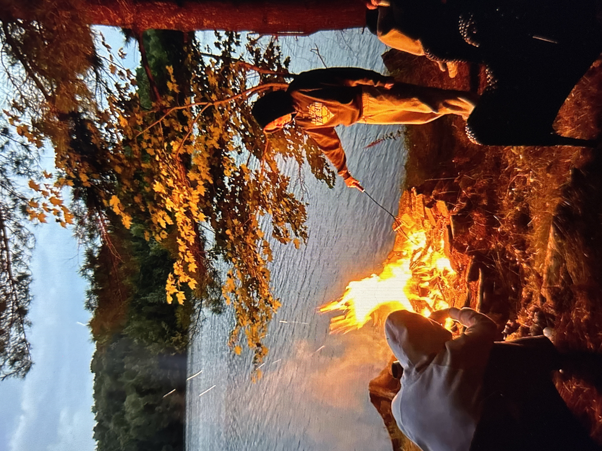 The media file depicts a group of people gathered around a campfire near a body of water, likely during the evening. The scene is set in a natural environment with trees and foliage visible. A prominent bonfire is burning, casting a warm glow on the surroundings and illuminating the faces of those present. One individual, wearing a hooded sweatshirt and a beanie, is seen holding a stick over the flames, possibly to roast marshmallows or food. Another person, dressed in a white hoodie, is partially visible in the foreground, their back turned to the camera. The water behind them appears calm, reflecting the light from the fire and the sky. The time of day is suggested to be twilight or early evening, given the warm, soft lighting and the presence of a campfire. The overall atmosphere seems relaxed and convivial, indicative of a camping or outdoor gathering. The image resolution is moderate, with some visual noise, and the orientation is rotated. No visible text is present in the image. The location context of Pastavy, Belarus, suggests a temperate climate and a natural landscape typical of the region.