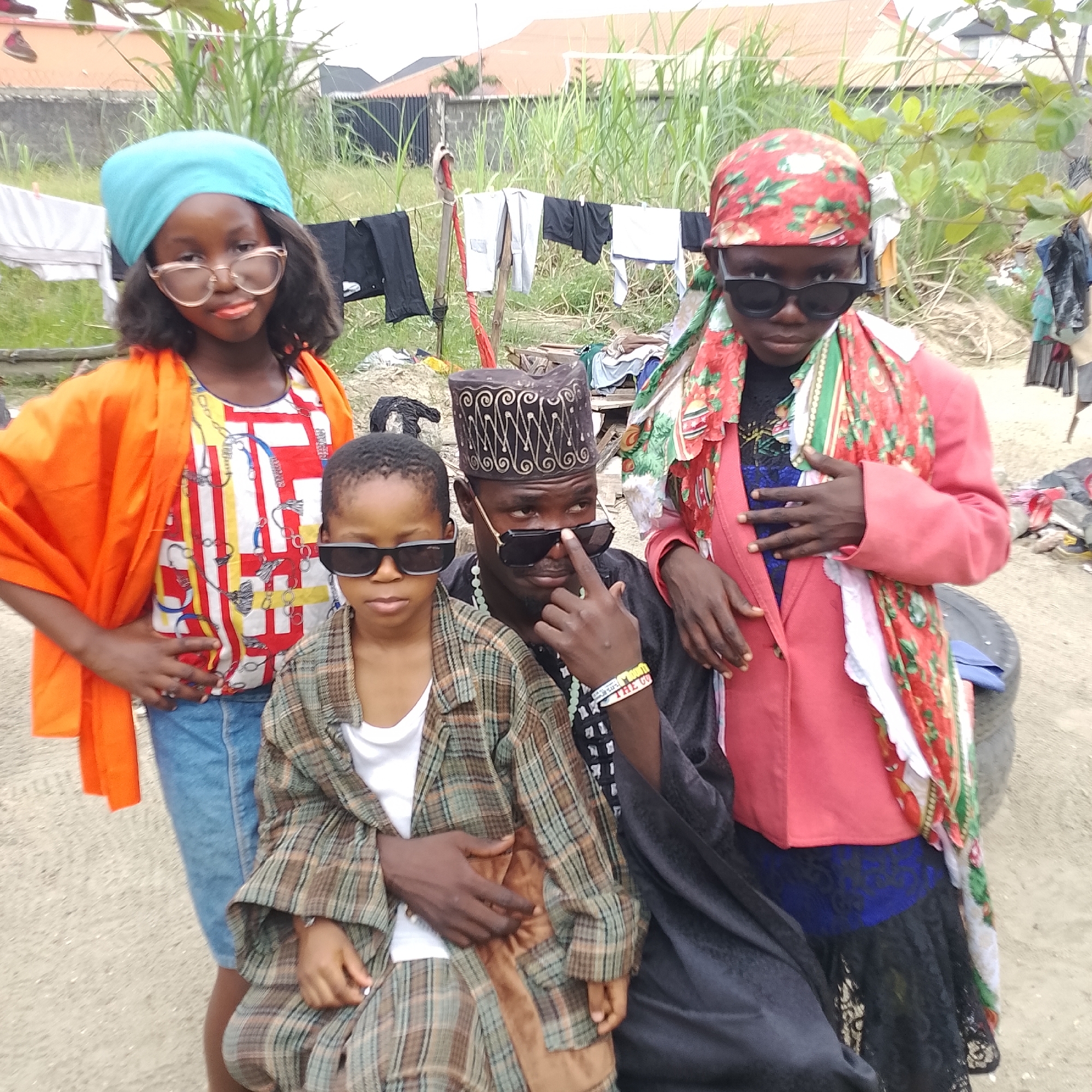 In the city of Ado, Nigeria, an outdoor scene unfolds. Three children and an adult male are positioned in the foreground, with lush green vegetation and residential buildings in the background. The adult male, wearing a traditional cap and dark attire, is seated, with a child on his lap and another standing beside him. A third child stands to his left. All three children and the adult are wearing sunglasses, adding a playful element to the scene. The clothing of the children is vibrant and diverse, featuring colorful tops and headscarves. Laundry hangs on a line behind the group, suggesting a domestic setting. The ground is covered with sand and scattered debris, hinting at a casual, perhaps informal, environment.