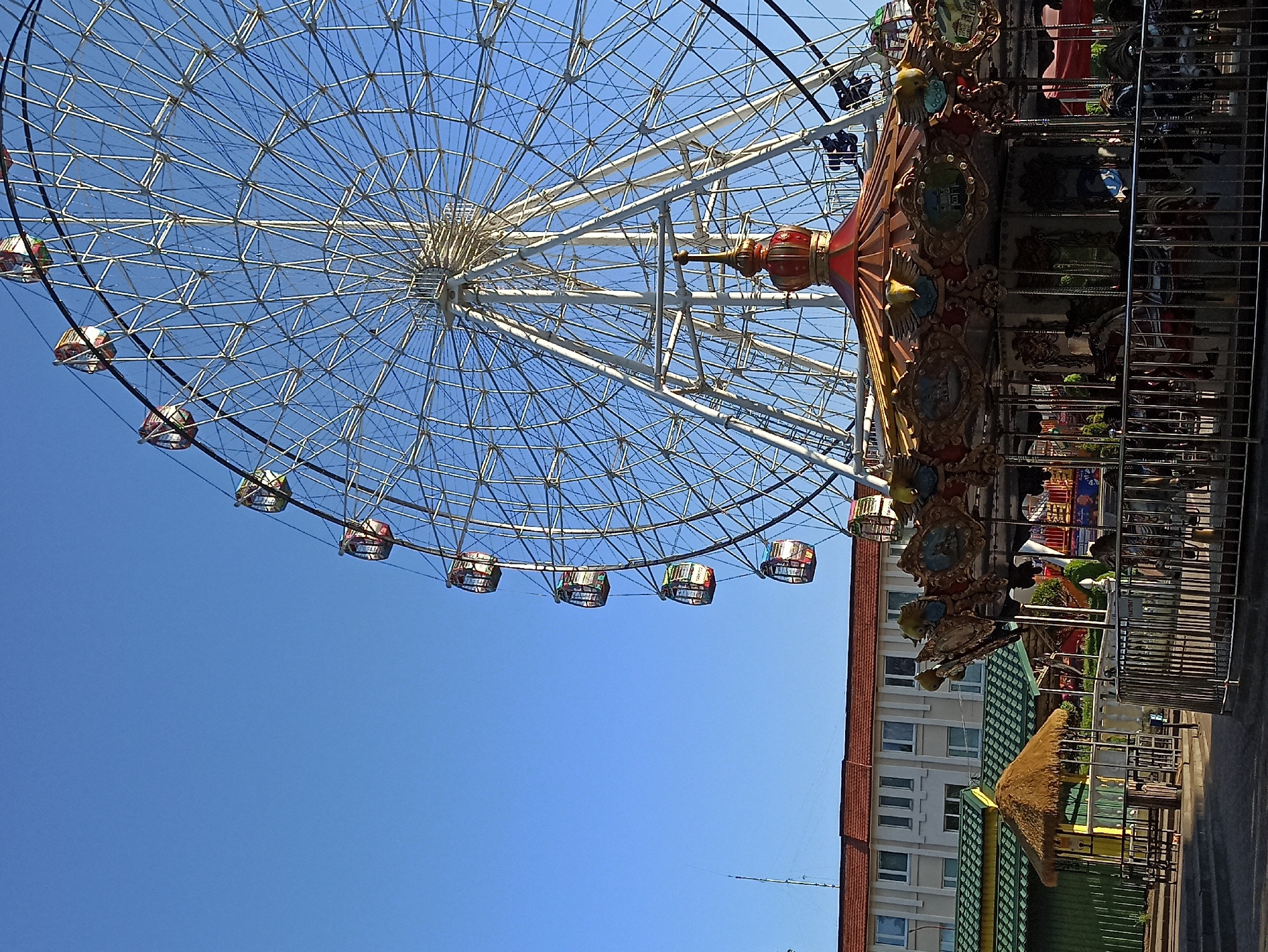 The image shows a Ferris wheel and a carousel in Hrodna, Belarus. The Ferris wheel is white with colorful passenger cars. The carousel is ornately decorated with carvings and lights. The sky is clear blue. A building is visible in the background, beyond the rides. No people are visible. The camera angle looks up at the Ferris wheel and the carousel is to the right of the wheel.