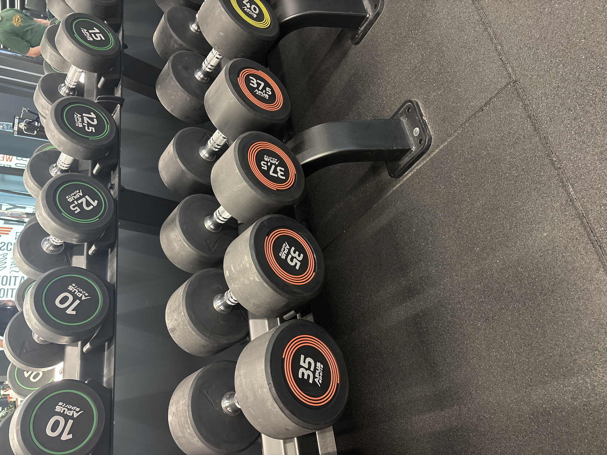 The image shows a rack of dumbbells in a gym. The dumbbells range in weight from 10 to 40 kilograms and are arranged in ascending order. The dumbbells have a black rubber coating with the weight displayed in a contrasting color (green, yellow or orange) with the words "APUS Sports" on them. The rack is made of black metal. The floor is covered in black rubber mats. The picture may be taken in a gym. The location is unknown and cannot be determined from the image.