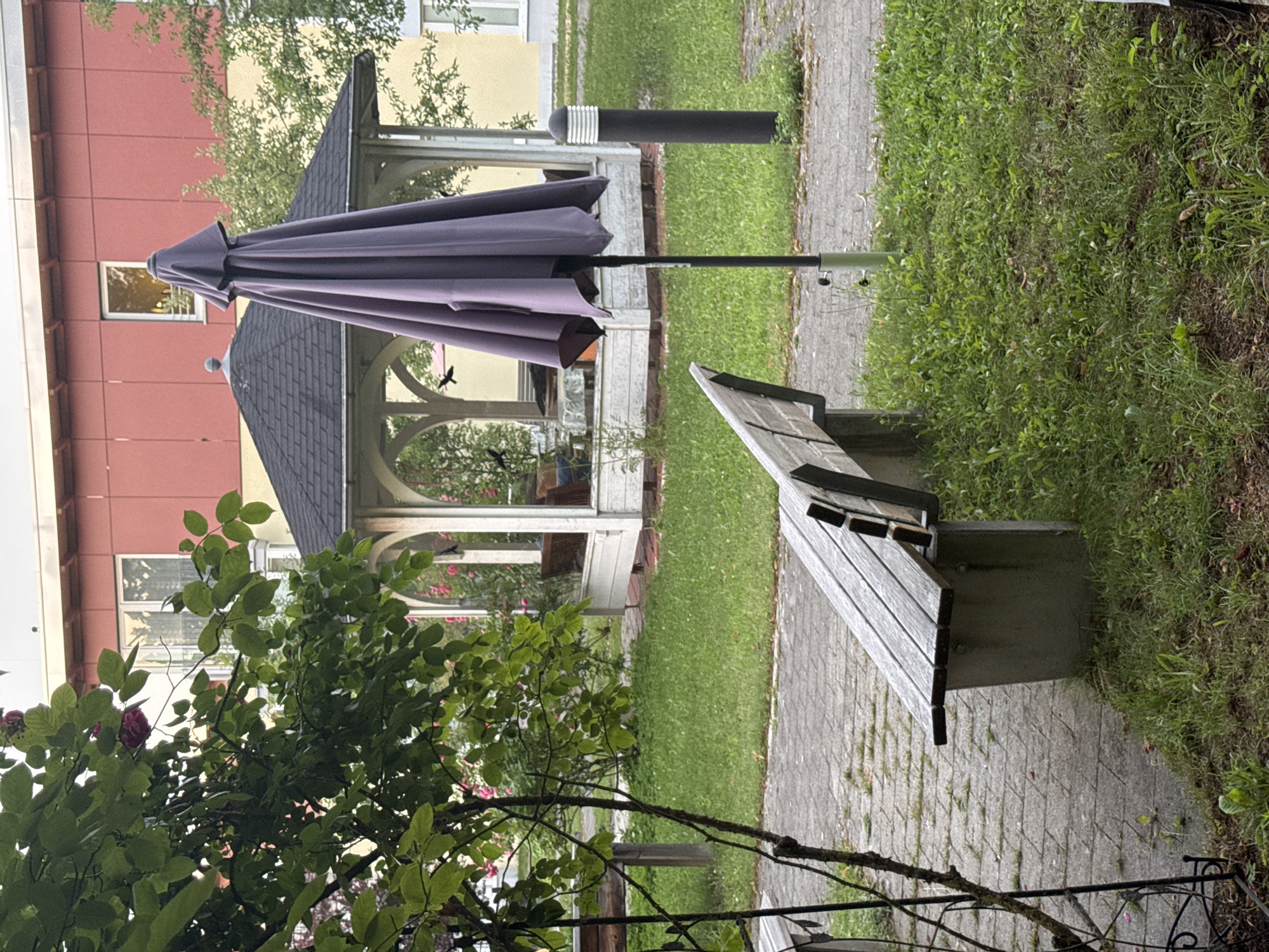 The image depicts a tranquil outdoor scene, likely in Höhenkirchen-Siegertsbrunn, Germany, featuring a paved pathway and a lush green lawn. In the foreground, a weathered wooden bench with concrete supports rests on the pathway, angled towards the right. To the left of the bench, a young tree with thin branches and green leaves is visible, with some pink flowers blooming amongst the foliage.
Further into the scene, a large, closed, grey umbrella stands prominently on a metal pole, casting a shadow on the grass. Behind the umbrella, a white gazebo with a dark, multi-faceted roof is situated. Inside the gazebo, a round table is covered with a clear plastic sheet, suggesting it's set for use or protection. Several dark silhouettes, possibly birds, are depicted in flight within or near the gazebo.
In the background, a building with a red facade and lighter colored trim rises above the gazebo. The building has visible windows, some with bay window extensions. The overall impression is one of a well-maintained public space, possibly a park or communal garden area within the town. The lighting suggests it is daytime, with soft light filtering through the trees and the sky is overcast. There are no people visible in the image.