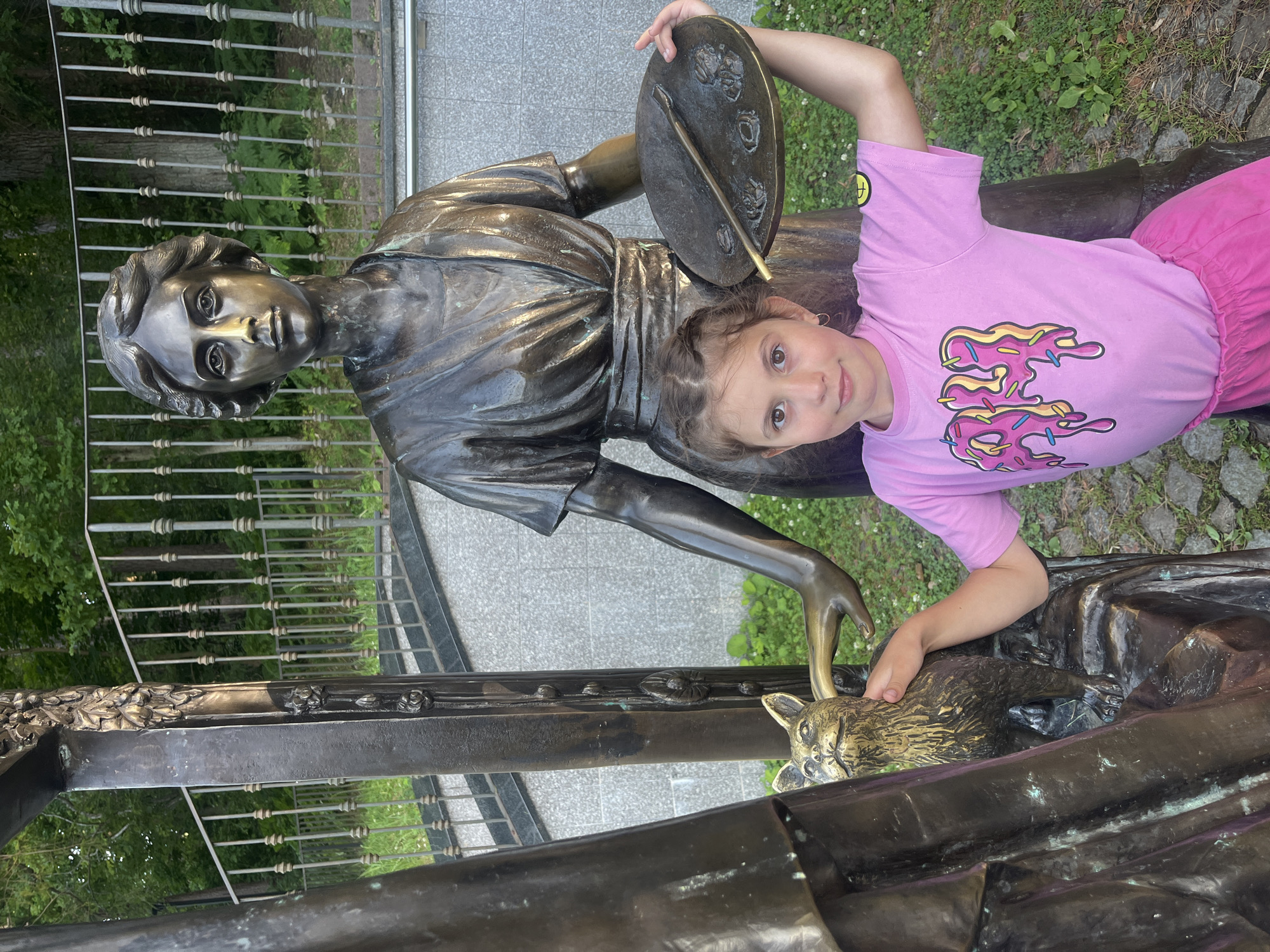 A young girl with brown hair, wearing a pink t-shirt with a donut graphic and pink shorts, stands next to a bronze sculpture of a woman holding a palette and brush, and a bronze cat. The girl is touching the bronze cat. The sculpture is located in Svetlogorsk, Russia, with a fence and trees in the background. The scene appears to be outdoors.