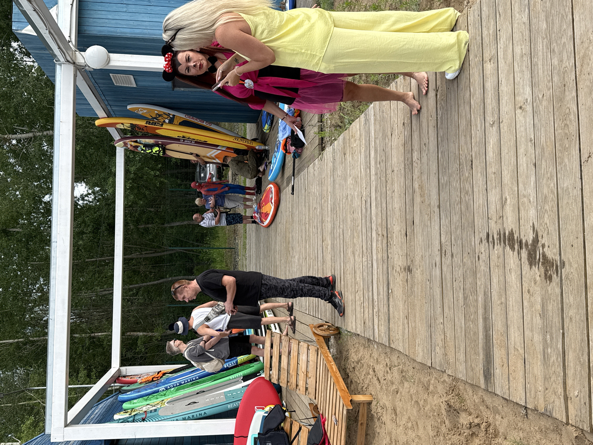 An outdoor scene on a wooden boardwalk near a body of water, likely a beach or lakefront, in Chaikovsky, Russia. Several people are present, engaged in various activities. In the foreground, two women are interacting, one wearing a yellow jumpsuit and the other a pink dress and Minnie Mouse ears, both looking at a phone. To the left, three men stand near a rack of paddleboards. A wooden lounge chair sits on the sand. In the background, other individuals are visible, including one person dressed in a Spider-Man costume, and a blue wooden building with a white structure overhead. The environment includes sandy ground, a wooden boardwalk, and a dense forest in the background.