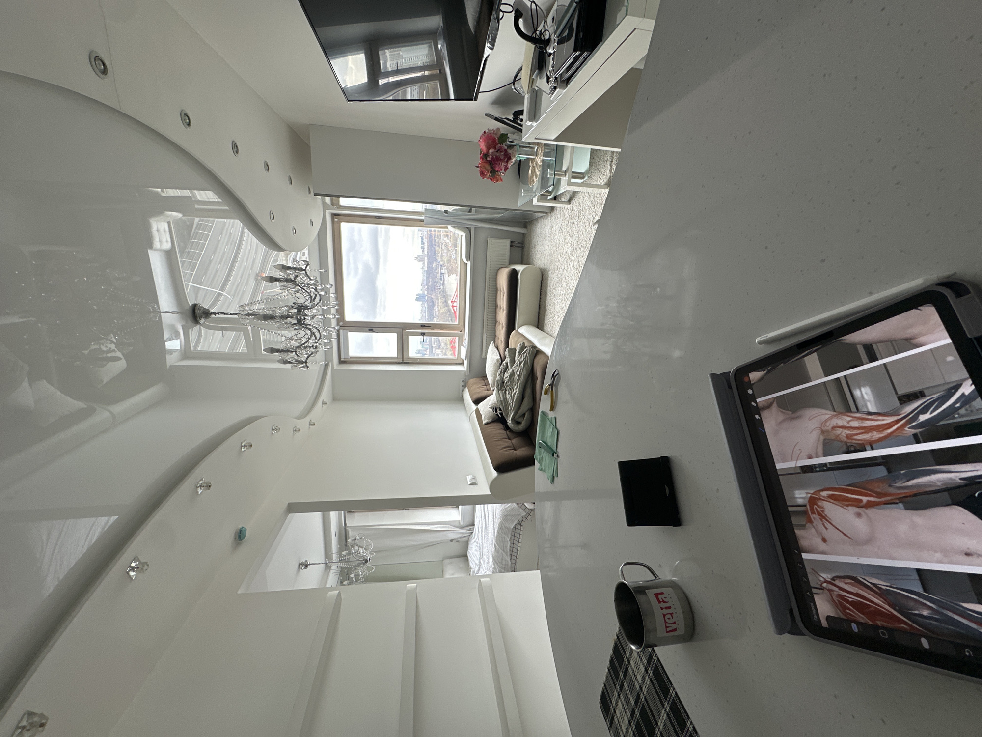 The image shows a modern, upscale apartment interior. The primary focus is on a bright, white kitchen counter where an iPad displays anatomical drawings. A mug and a notebook sit beside the iPad. In the background, through a large window, a cityscape is visible. The view is consistent with a high-rise location in Moscow, Russia, looking towards the city skyline. Further into the apartment, a living area with a sofa and a bedroom area can be seen. The overall aesthetic is modern and minimalist.