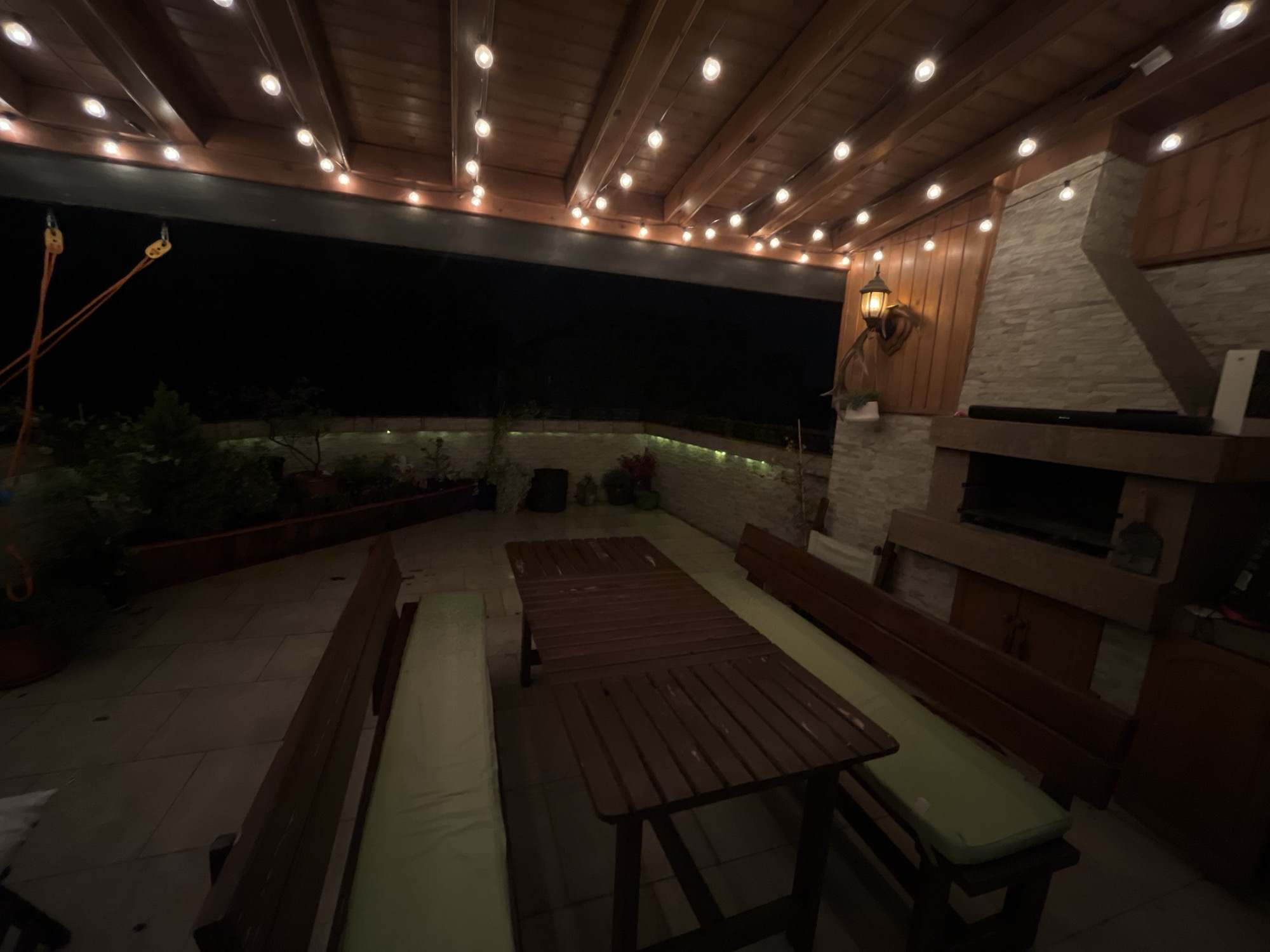 This image depicts an outdoor patio area at night in Plovdiv, Bulgaria, illuminated by numerous overhead string lights and a wall-mounted lantern. The patio features a dark wooden slatted pergola roof structure, from which the string lights are suspended. On the left side, two orange ropes or cables hang from the roof structure, each terminating in a small yellow pulley or attachment mechanism against a dark background of the night sky and distant foliage. In the center, a long, rectangular wooden table with a slatted surface is flanked by two dark wooden benches, each fitted with a light green cushion. The patio floor is paved with light-colored square tiles. Along the right side of the image, a prominent stone-clad structure, likely a fireplace or barbecue, rises towards the pergola. It features a large opening for grilling at its base. To the left of this stone structure, a wall-mounted lantern with a warm glow illuminates a decorative deer head with antlers positioned below it, affixed to a wooden wall panel. On a wooden ledge above the grill opening, a dark soundbar and a white rectangular object (possibly a speaker) are visible. Further to the right, a dark wooden cabinet or storage unit is partially visible. Behind the table and benches, a raised planter or low wall runs across the back, containing various plants and shrubs, subtly illuminated by ground lights or smaller string lights. No individuals or animals are present in the scene.