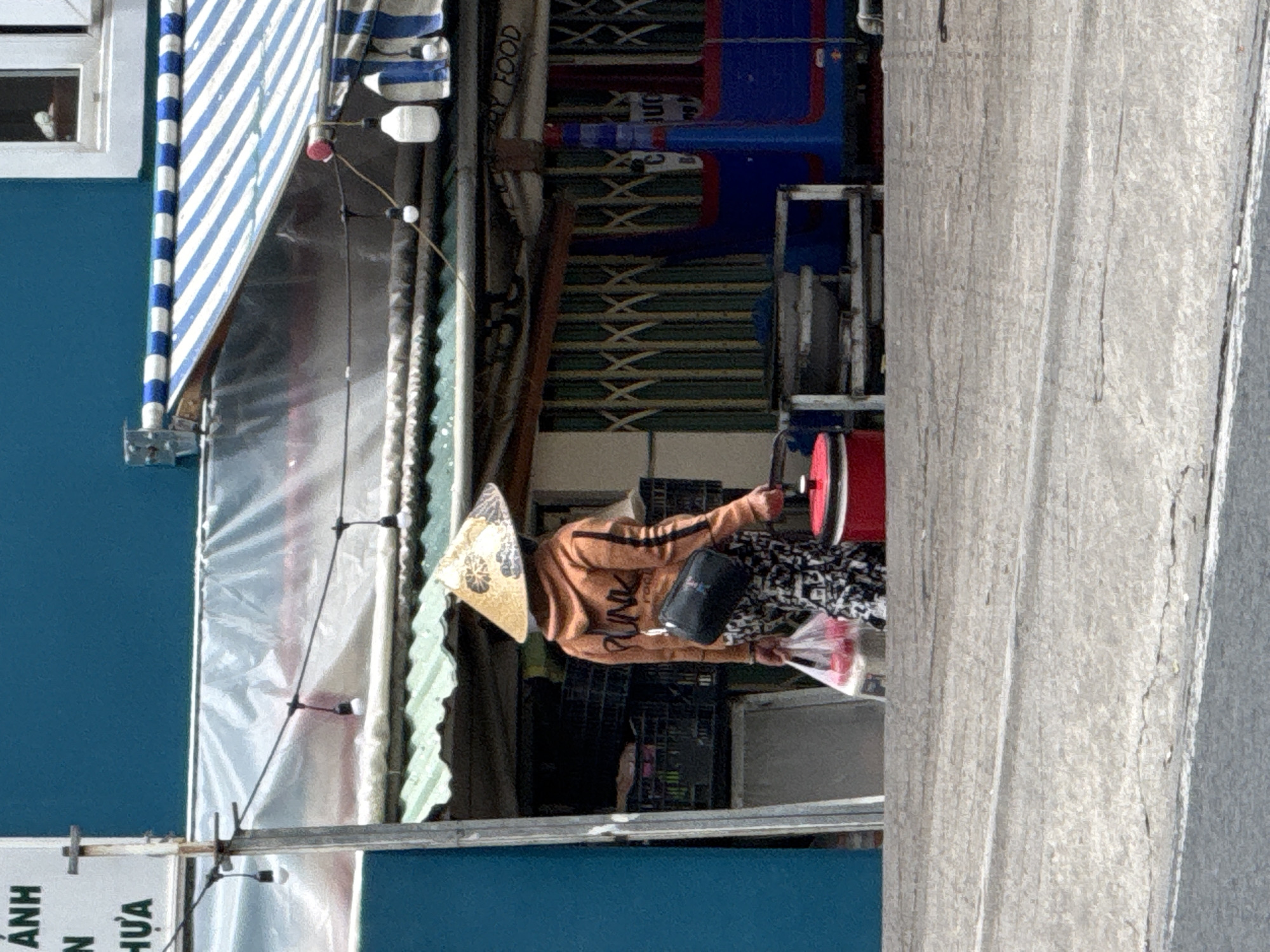 A person wearing a conical hat and an orange hoodie is standing on a street in Da Lat, Vietnam, carrying shopping bags and a red cooking pot. The person is facing away from the camera and is dressed in patterned pants. Behind them, a stall with a green awning is visible, and a blue building serves as the backdrop. The street appears to be paved with concrete. The lighting suggests it is daytime.