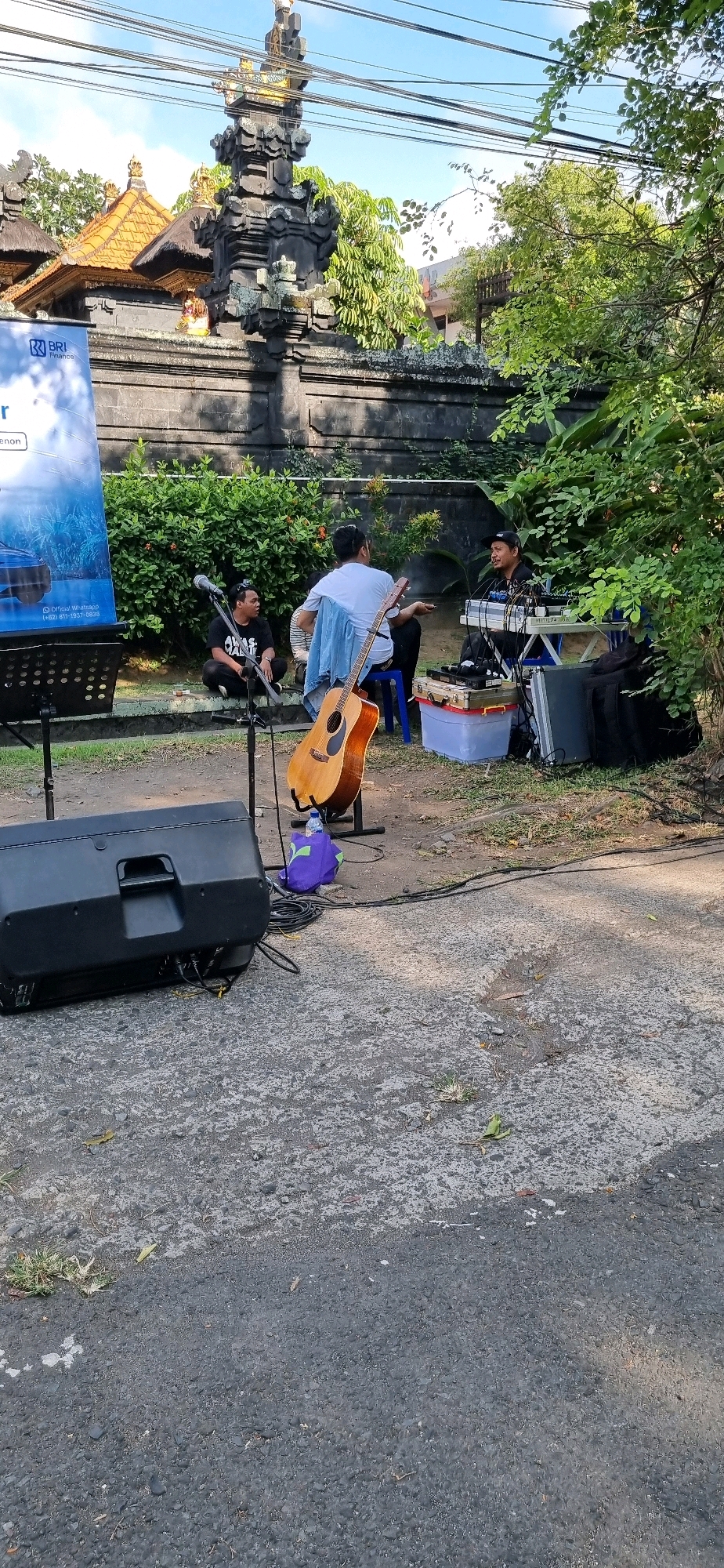 Three individuals are seated outdoors, appearing to be a musical trio, with a prominent religious structure in the background. To the left, a large banner with text and a blue logo, possibly indicating a financial institution, is erected on a stand. In the foreground, a large black speaker is positioned on the ground. In the middle ground, one person sits on the ground, facing left, with a microphone stand in front of them. Another individual is seated on a blue chair, facing right, holding a guitar. A third person is seated behind a table or console, operating what appears to be musical equipment. An acoustic guitar stands upright in front of the person on the chair. The setting appears to be a public space with paved ground and patches of grass, bordered by a stone wall and lush greenery. The architecture in the background, characterized by intricate stone carvings and tiered roofs, is indicative of a temple or a traditional Balinese structure. This scene could be situated in a public area of Denpasar, Indonesia, given the architectural style and the presence of a financial institution banner, suggesting a commercial or cultural event.