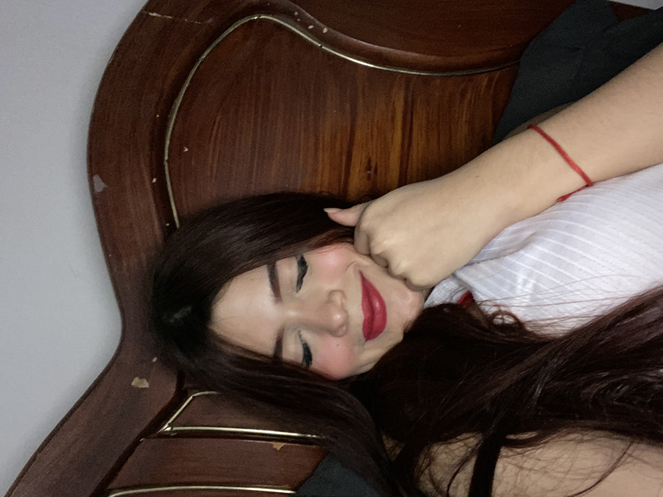 A woman is lying down with her face tilted to the side. Her long, dark brown hair cascades around her face. She is wearing red lipstick and her eyes are closed. Her left hand is near her cheek. She is wearing a white striped shirt and a red string bracelet. The background features a wooden headboard, with some wear and tear visible, and a light gray wall.