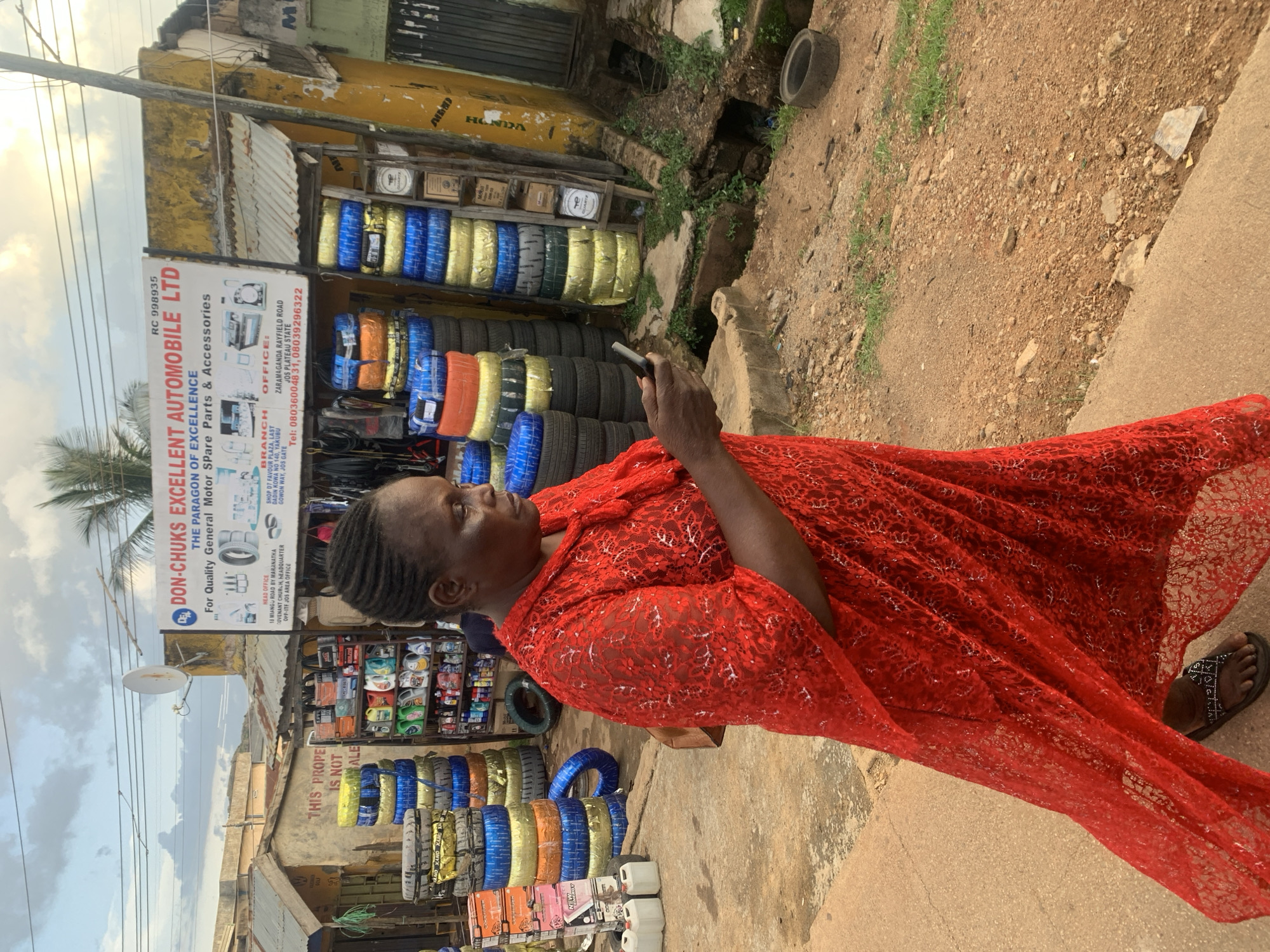This is a vertical, outdoor photograph taken in Jos, Nigeria, likely during the day, as indicated by the bright lighting and cloudy sky. The primary subject is a woman dressed in a vibrant red lace dress, standing in front of a shop. She is holding a smartphone in her right hand and appears to be looking towards the left of the frame. Her expression is neutral or slightly thoughtful.
The setting is a bustling roadside area, typical of many markets or commercial streets in Nigeria. Behind the woman, a shop displays a large quantity of tires, stacked in various colors including blue, yellow, and black. Some of the tires are individually wrapped. The shop's facade is painted yellow and shows some wear and tear. A prominent sign for "DON-CHUKS EXCELLENT AUTOMOBILE LTD" is visible, providing contact information and listing the business as a provider of quality general motor spare parts and accessories. Other items, like bottles of what appear to be engine oil or lubricants, are also displayed on shelves.
To the right of the shop and the woman, the ground is rough and dusty, with some small stones and sparse green vegetation growing. A discarded tire is visible on the ground, and a concrete gutter runs along the edge of the dirt path. Further in the background, there are more buildings and structures, suggesting a densely populated commercial area. The overall atmosphere appears to be that of a typical Nigerian roadside market.
