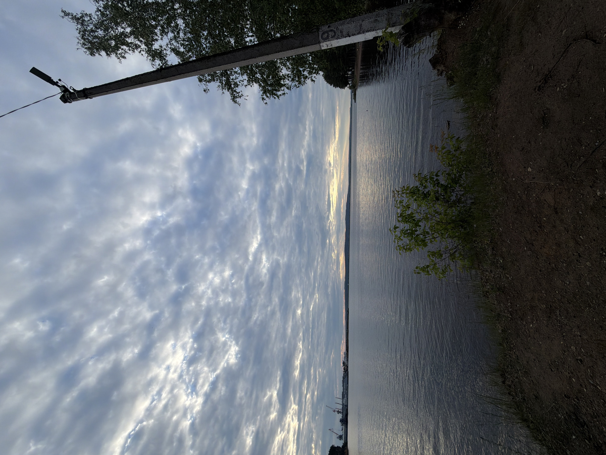 A wide shot captures a serene lake scene at dusk, with a cloudy sky and a distant industrial port. The foreground features a dirt embankment with some greenery, and a concrete utility pole stands to the right, adorned with a number "9." In the distance, a line of trees borders the opposite shore, and the port's cranes are visible. The water is calm, reflecting the subdued light of the setting sun. The location is the Kama River in Chaikovsky, Russia.