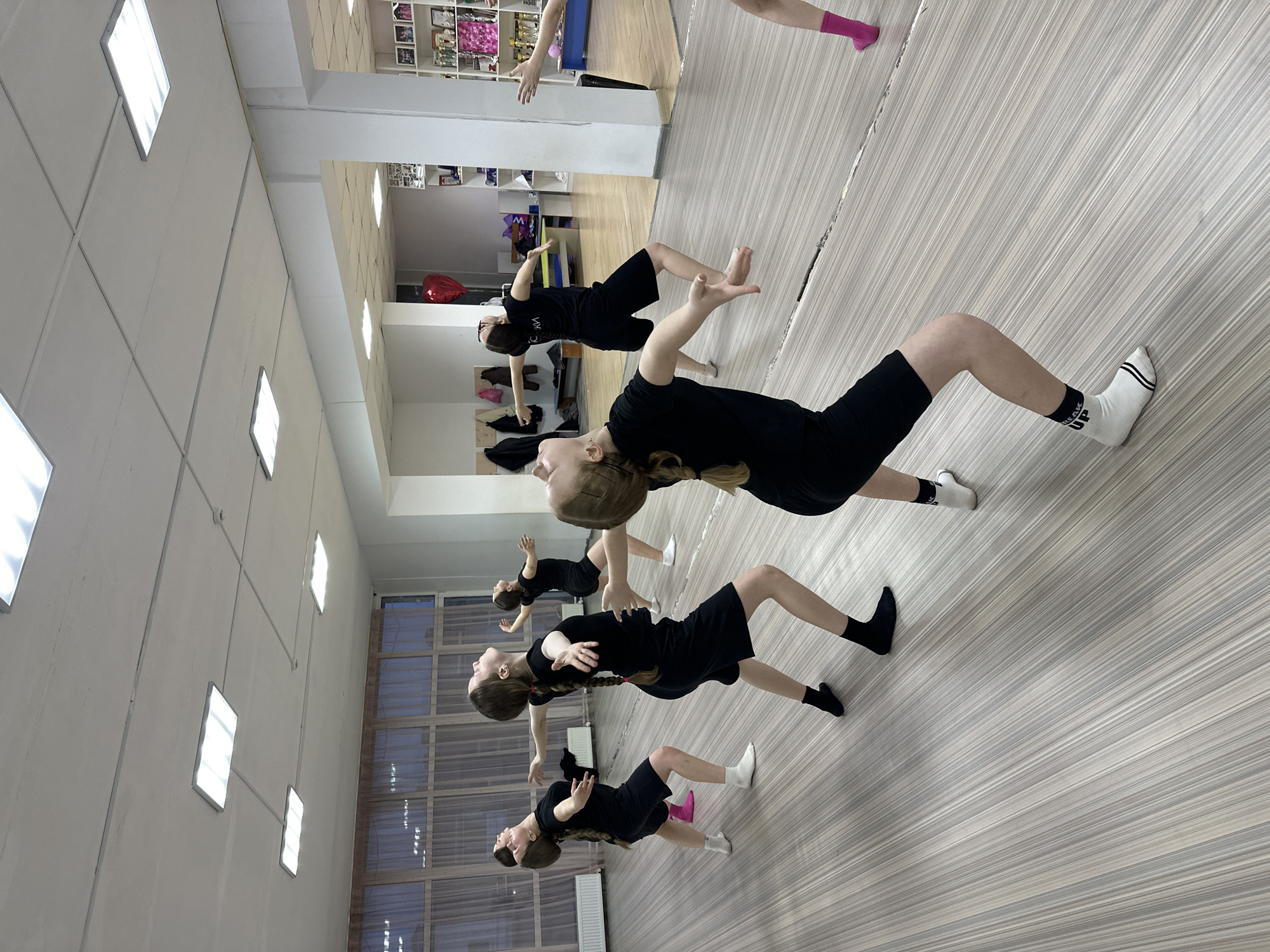 A group of young people, primarily girls, are performing a synchronized dance or exercise routine in an indoor studio. They are uniformly dressed in black t-shirts and black shorts or skirts, with their hair mostly styled in braids or ponytails.
The individuals are captured in a moment of a back-arching movement, leaning backward with their arms extended out or slightly upward, and their heads tilted back, looking towards the ceiling. Their expressions appear focused and concentrated. Some are wearing black socks, while others have white socks; one person is seen with a single pink sock. A white sock on one participant's foot shows partially visible text, possibly "CREATE".
The setting is a well-lit studio with light-colored, possibly wooden or laminate, flooring. The ceiling is fitted with numerous square fluorescent light panels. One wall features a large mirror, reflecting the participants and a background area with shelves displaying trophies and other items, suggesting a dance school or training facility. Another wall has large windows, offering an indistinct view of the outside. The scene conveys an atmosphere of dedicated practice during a dance or fitness session.