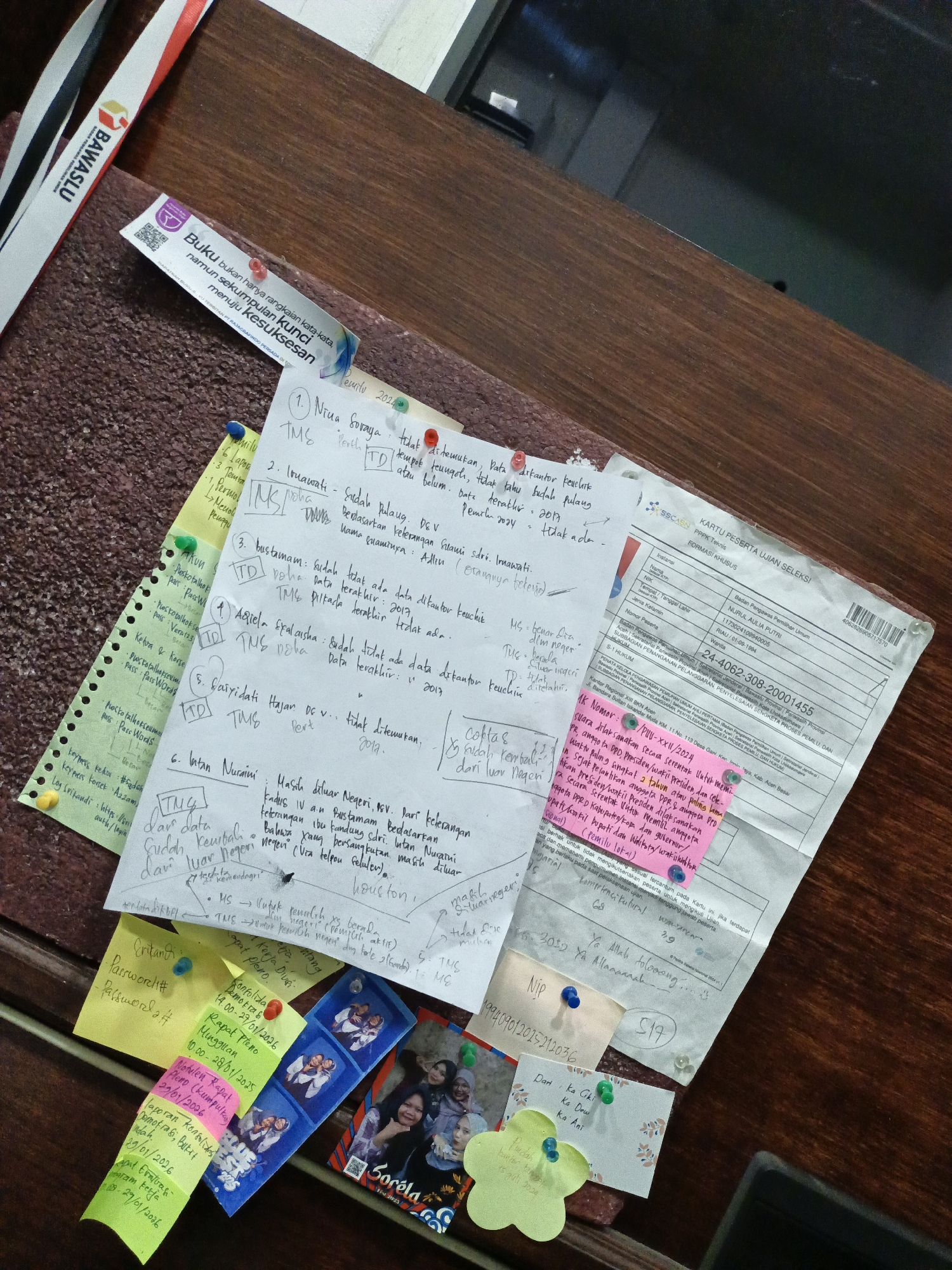 A desk is cluttered with papers, sticky notes, photos, and other items.
Prominently visible is a "KARTU PESERTA UJIAN SELEKSI" (Examination Selection Participant Card) with the name NURUL AULIA PUTRI and details related to the Badan Pengawas Pemilihan Umum (Election Supervisory Body). A number of handwritten notes on paper are pinned to the surface. One note references the city of Lhokseumawe in Indonesia. Other handwritten notes appear to be reports or records, including the acronyms TMS and TD. Sticky notes contain passwords and dates. A strip of photos shows a group of young women and the word "Sorela." A tag is attached to a lanyard and has the word "BAWASLU" printed on it. There are blue and yellow pushpins scattered around the desktop.
