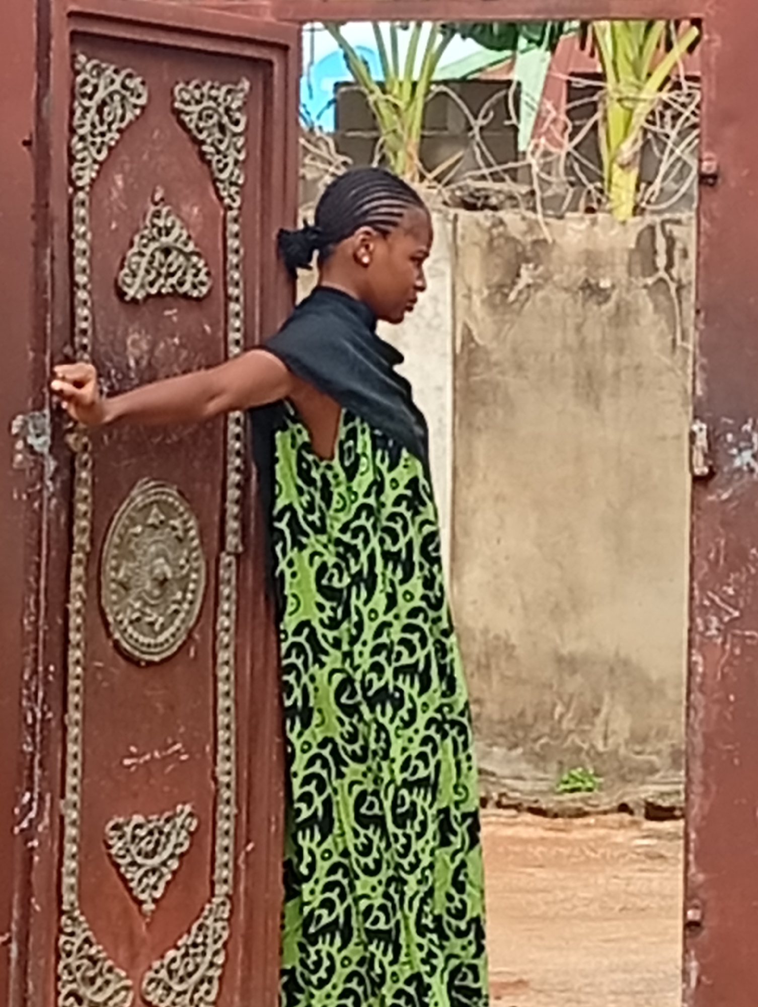 A person is standing in a doorway. The person is wearing a green and black patterned dress and a black shawl. The doorway is ornate and made of reddish-brown material. Behind the doorway is a concrete wall topped with barbed wire and vegetation. The location is in the city of Unknown, Unknown.