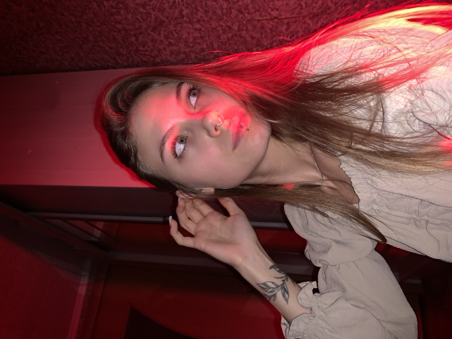 The image depicts a young female positioned indoors under prominent red lighting. Her head is turned to her right, with her gaze directed towards the viewer's right. Her left hand is raised, with fingers near her left ear. She has long, light brown hair. Distinctive features include a silver hoop septum piercing and two small stud piercings below her lower lip. A tattoo of a leafy branch is visible on her left forearm. She is wearing a light-colored, likely beige, ruffled top and a thin silver chain necklace. The background on the viewer's right features a textured wall heavily saturated with red light, while the left side of the background is darker and less defined. The scene is located in the city of Фоки, Russia.