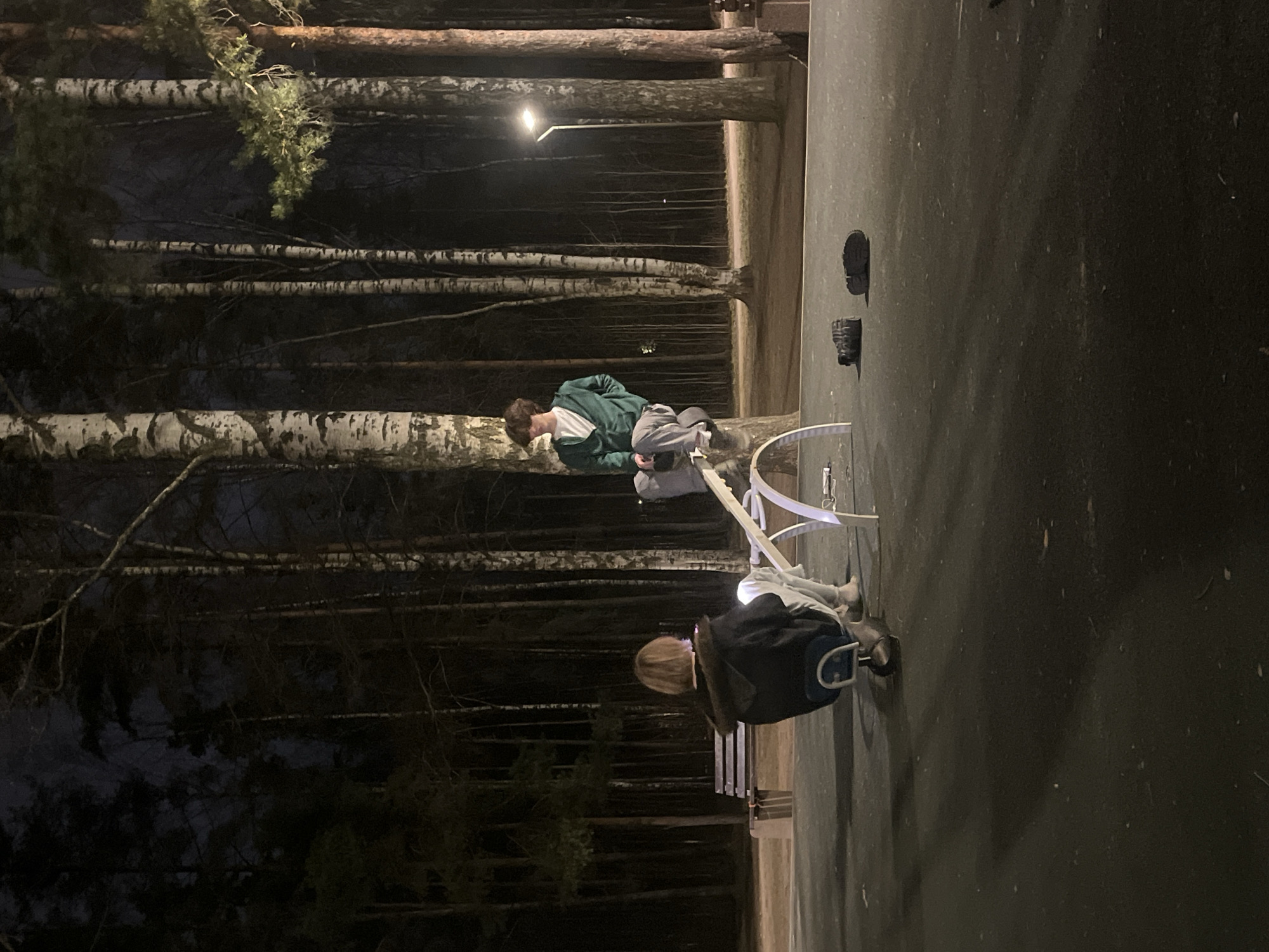 The image depicts two people sitting on a park bench in what appears to be a wooded area. The location is in Saint Petersburg, Russia, judging by the prevalence of birch trees and general atmosphere of the landscape. One person, wearing a green jacket and gray pants, is seated upright on the bench. The second person, wearing a black jacket, is sitting beside them. A row of birch trees is visible in the background and a path or paved area stretches horizontally across the top part of the image. The light is dim, suggesting it is either dusk or nighttime. Two objects are seen attached to the vertical surface, possibly a wall, to the right of the bench; it appears to be a shoe and a hat.