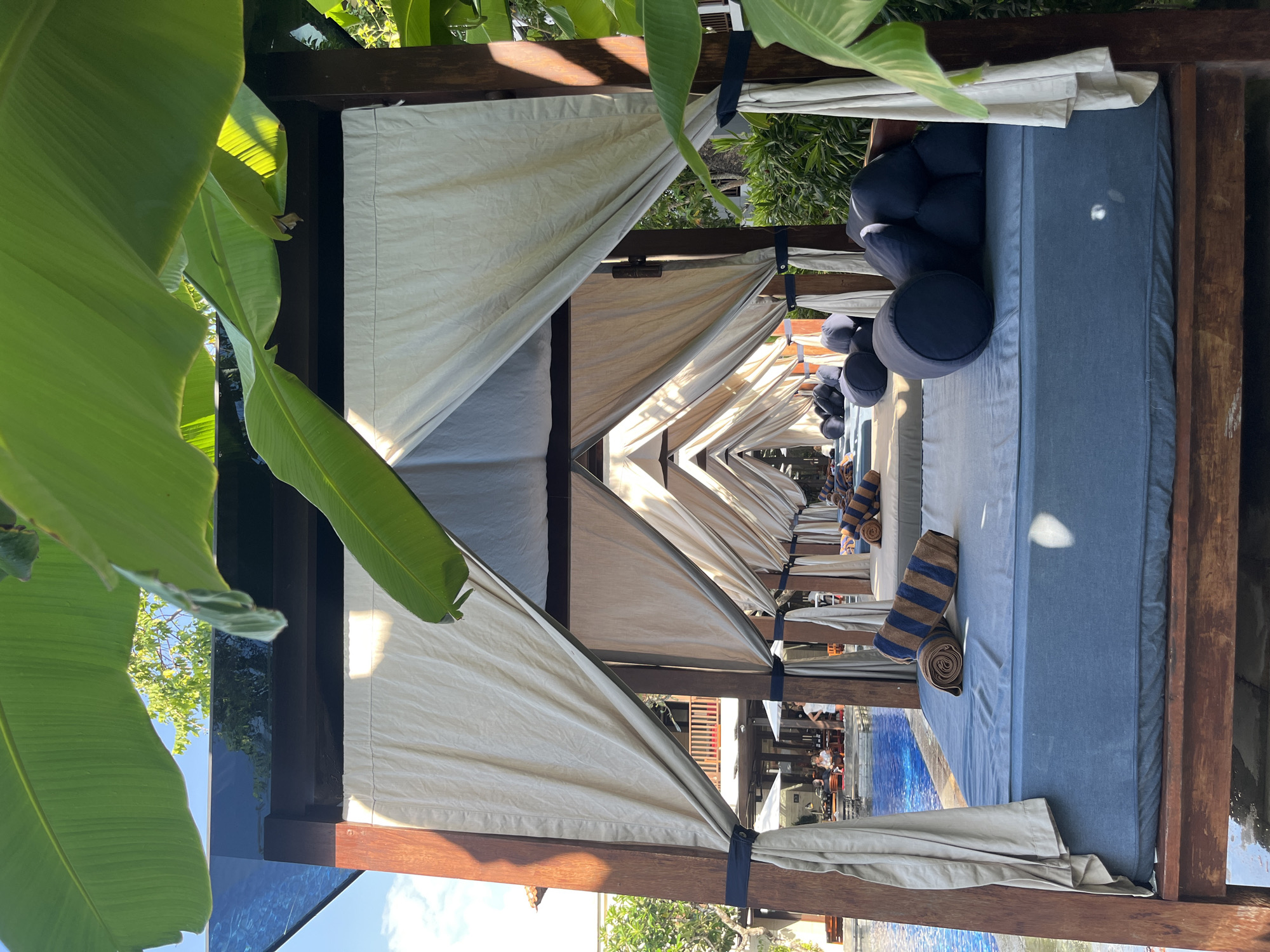 The image captures a serene poolside scene at the Anantara Uluwatu Bali Resort in Kuta Selatan, Indonesia. In the foreground, a plush blue daybed is set within a wooden cabana, adorned with rolled towels featuring blue and brown stripes. The cabana is draped with light-colored fabric curtains, creating a sense of privacy and shade.
Beyond the immediate cabana, a series of similar structures recede into the distance, forming a symmetrical pathway. Each daybed appears to be equipped with dark blue cushions. To the left, a sparkling blue swimming pool is visible, with lounge chairs and umbrellas further in the background. People are seated at tables by the pool, suggesting a leisurely atmosphere.
Lush green foliage, including large banana leaves, frames the top and right edges of the image, adding a tropical touch to the setting. The sunlight casts dappled shadows on the ground and across the cabanas, indicating it is daytime. The overall impression is one of relaxation and luxury, typical of a resort environment.