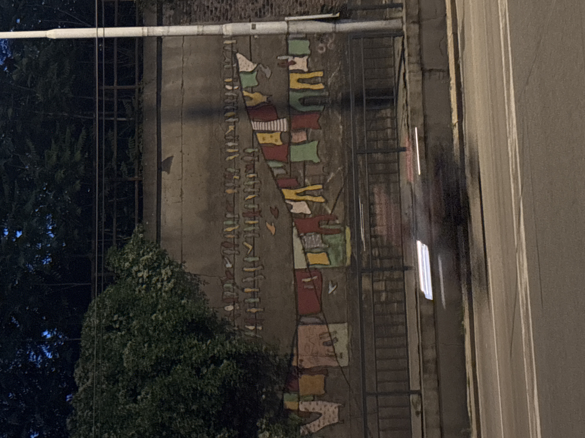 The image depicts a nighttime scene of a mural painted on a concrete wall in an urban environment. The mural features a stylized depiction of laundry hanging on a clothesline, with numerous items of clothing rendered in various colors. Above the clothesline, smaller figures resembling birds are painted. To the left of the mural, a large, leafy tree is visible, its branches extending towards the wall. A dark metal fence runs along the base of the wall. In the foreground, a road with visible lane markings is present, and a vehicle is blurred due to motion, indicated by streaks of light. A tall, white lamppost stands to the right of the mural. The overall lighting suggests it is nighttime, with artificial light sources illuminating parts of the scene. The setting appears to be outdoors, adjacent to a road.