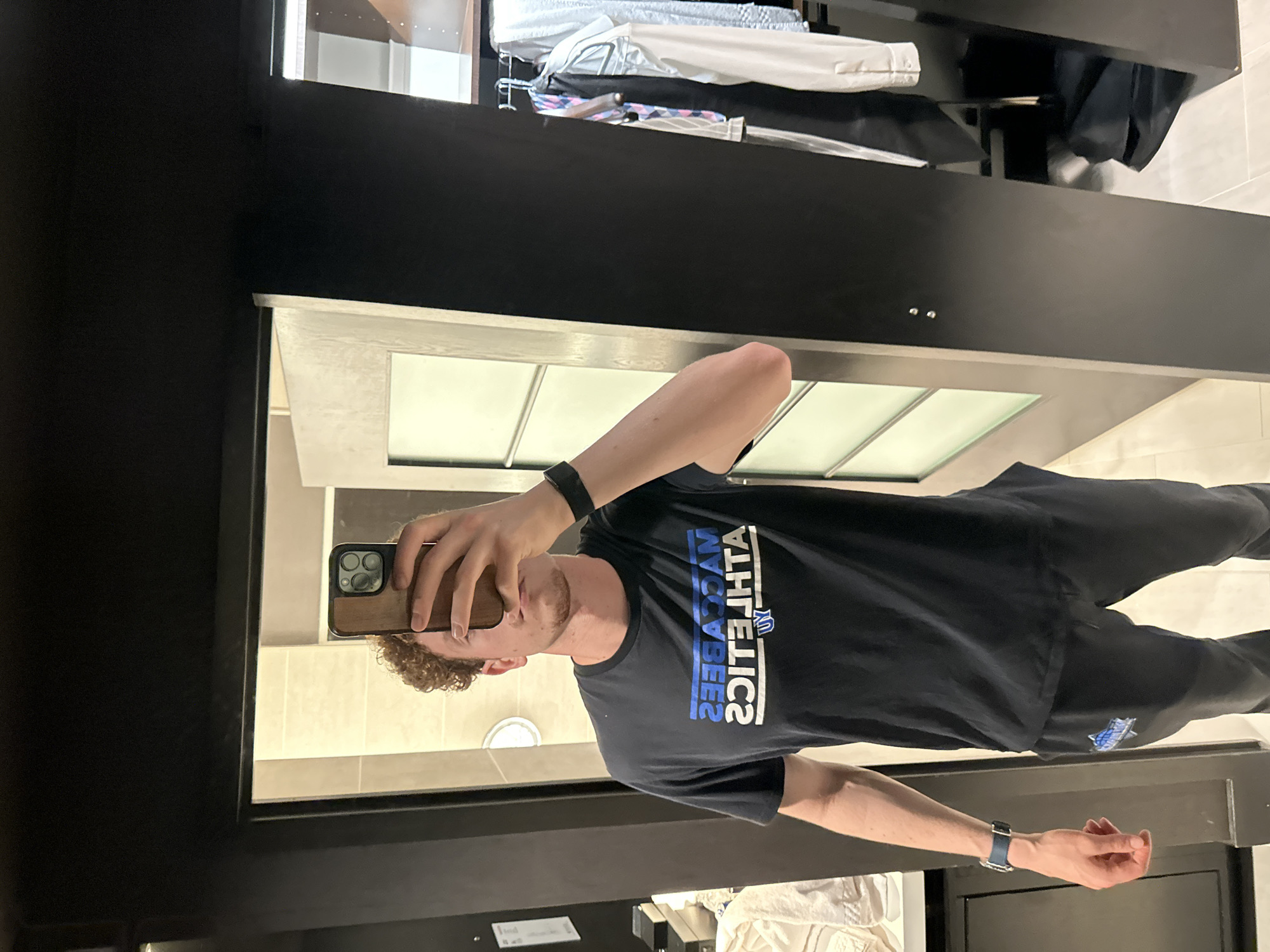 A man with curly brown hair is taking a selfie in a mirror. He is wearing a black t-shirt with "MACCABEES ATHLETICS" printed on it in blue and white, along with black pants and a black watch on his left wrist and a blue watch on his right. The mirror is framed in black, and the reflection shows a bathroom with beige tiles and cabinets above. Based on the shirt, the location is likely in College Park, United States of America, due to its association with Maccabees Athletics.