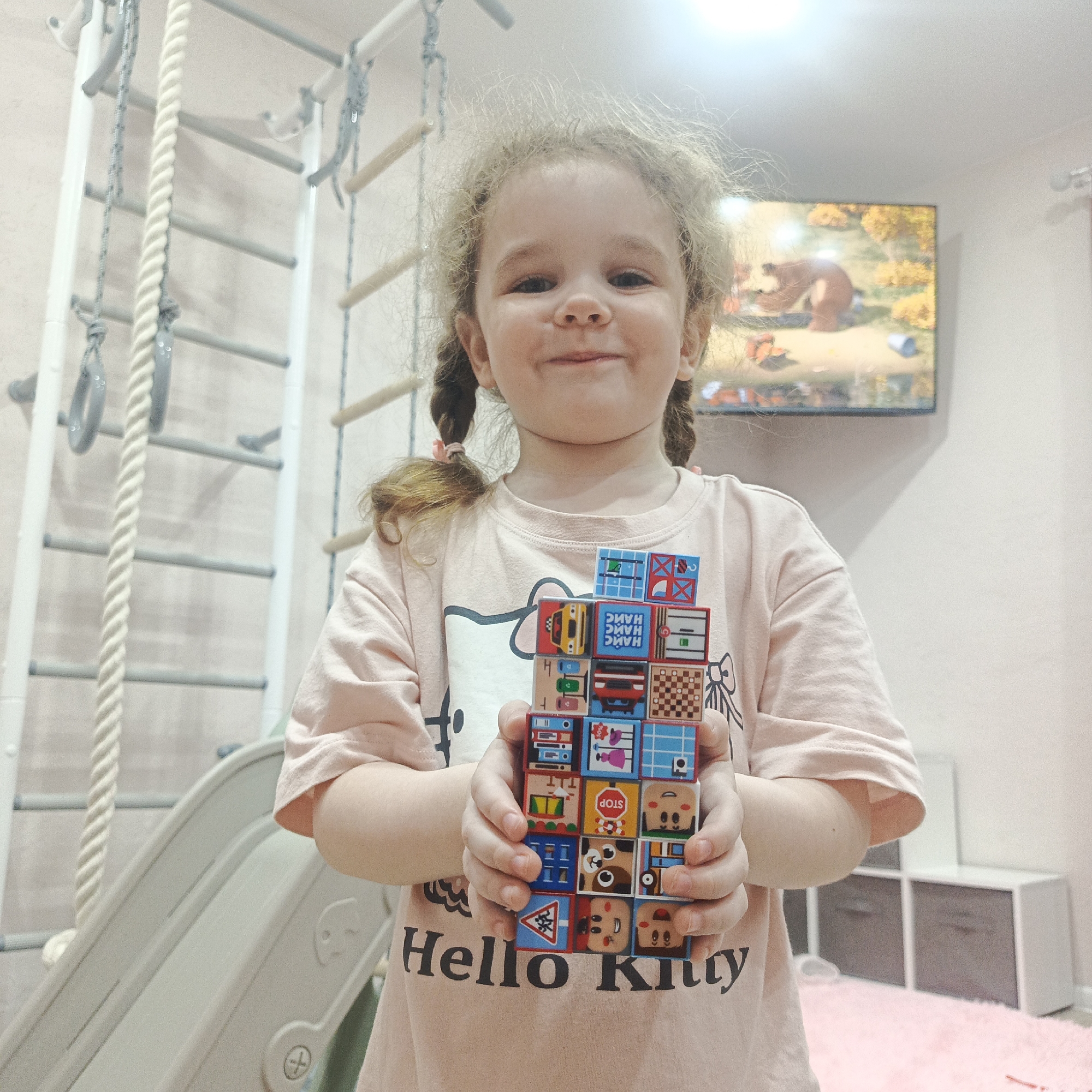 A young child stands indoors in Марковский, Russia, holding a stack of colorful blocks. The child has fair skin, curly blonde hair styled in pigtails, and is wearing a pink t-shirt with "Hello Kitty" printed on it. The blocks are decorated with various pictures of vehicles, faces, and symbols.
In the background, there is a climbing frame with ropes and rungs, as well as a plastic slide. A television is mounted on the wall, displaying an image of a bear. There are white storage units visible in the lower right of the frame. The overall environment appears to be a child's playroom.