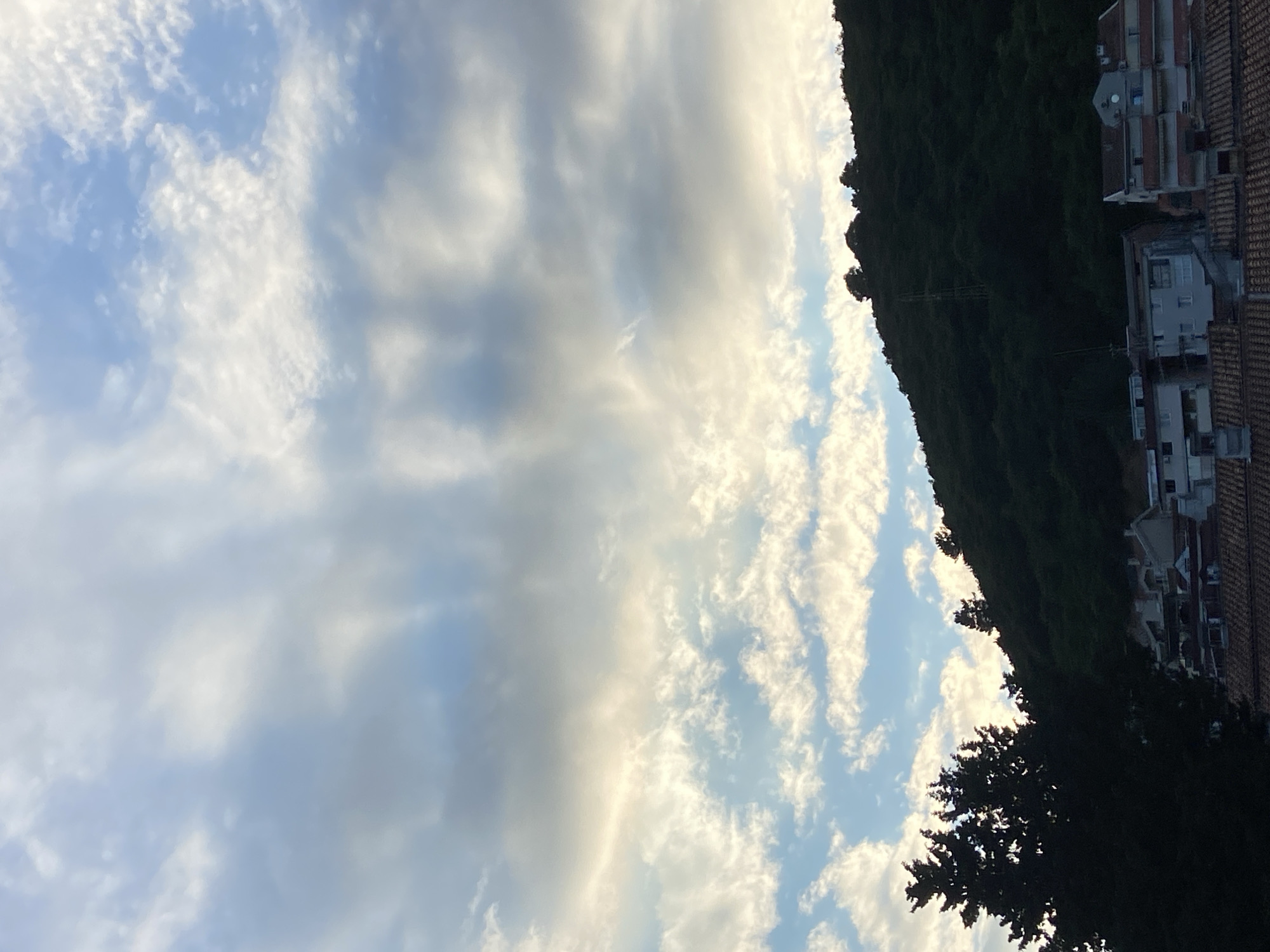 The image shows a view in Budva, Montenegro. The sky is filled with a mix of light blue and white clouds. On the right side of the image, there is a dark green, tree-covered hillside. Buildings are visible on the side of the hillside with tile roofs. Some trees are close.