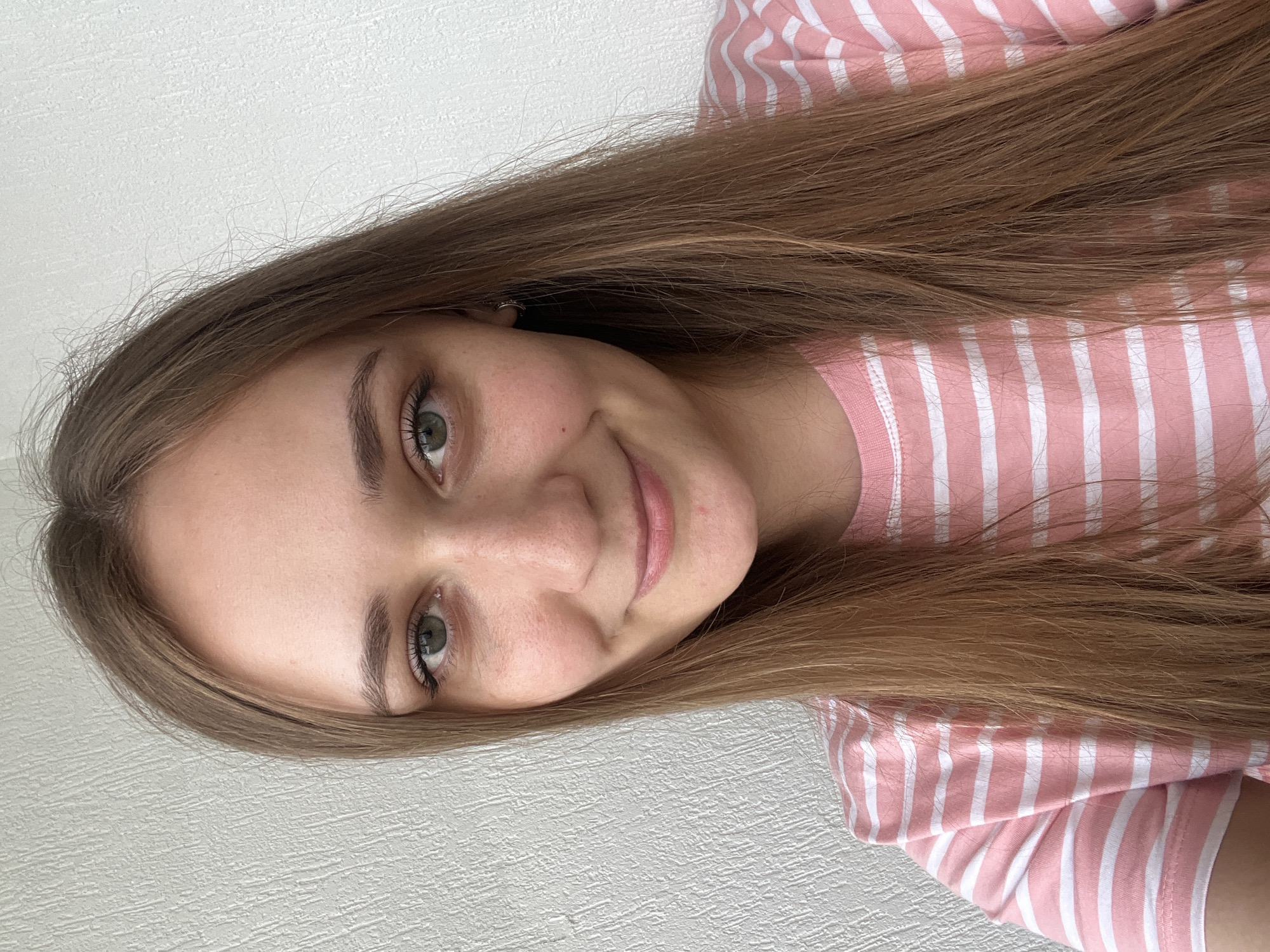 A woman is facing the camera, smiling gently. She has light skin, green eyes, and light brown hair. She is wearing a pink and white striped shirt. The background is a plain white wall. There is nothing that indicates the location is Yekaterinburg, Russia.