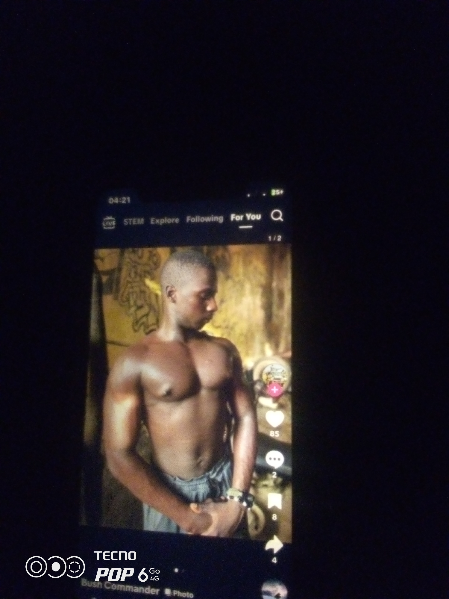 A medium shot captures a young Black man with short hair, wearing grey striped shorts, standing shirtless. His skin is dark and appears to be glistening with sweat, highlighting his muscular physique. He is facing slightly to his right, with his head tilted down. His left arm is bent at the elbow, with his hand near his waist, and his right arm is also bent, with his hand in a fist at his side. He is wearing beaded bracelets on his left wrist.
Behind him, a textured, light brown wall with some markings can be seen. The scene appears to be illuminated by artificial light, casting a warm glow on the subject. The image is displayed on a phone screen, as indicated by the interface elements visible at the top and bottom. These include a timestamp "04:21", navigation tabs "LIVE STEM Explore Following For You", a search icon, and the text "1/2". Social media icons, such as a heart with "85" likes, a speech bubble with "2" comments, and a bookmark icon with "8" shares, are present on the right side of the screen, suggesting the image is from a social media platform. At the bottom of the screen, "TECNO POP 6 Go 4G" is visible, indicating the phone model. The overall lighting of the image is dark, with the phone screen being the primary source of illumination. There is no discernible information about the specific location within Howayi, Nigeria, or any interaction between individuals in the image.