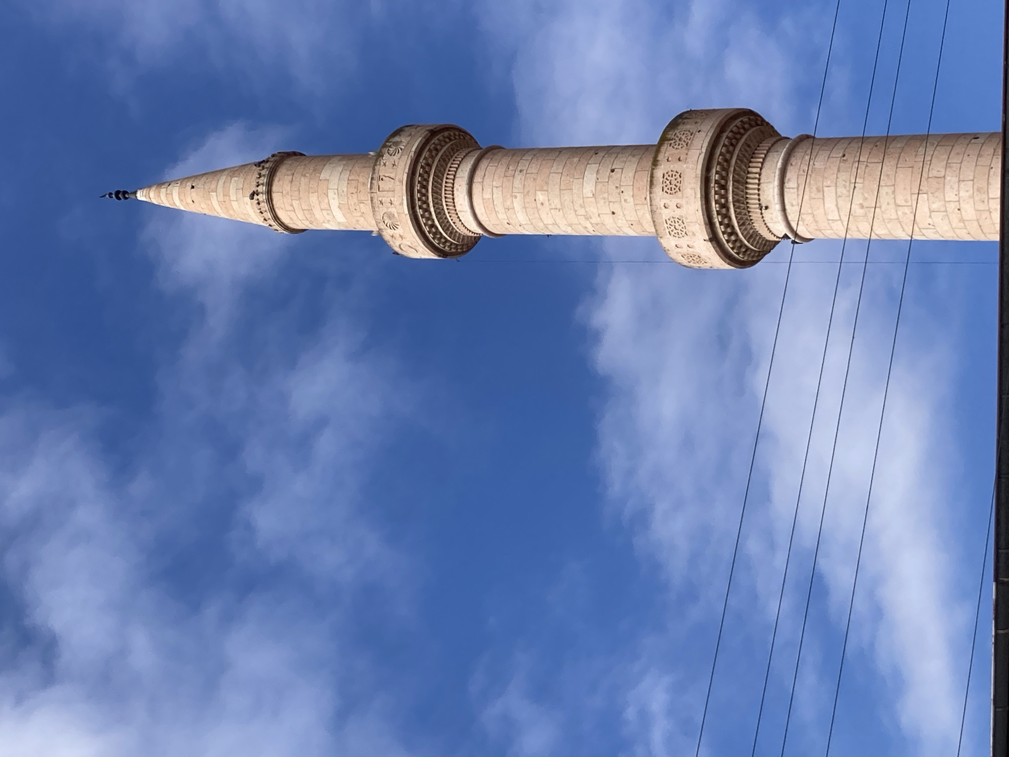 The image shows a close-up view of a tall, cylindrical minaret made of light-colored stone, tapering to a pointed spire. The minaret is adorned with decorative bands at different levels, featuring intricate carved patterns. The sky behind the minaret is a bright blue with scattered white clouds. Several parallel wires are visible in the foreground, running horizontally across the lower portion of the frame. The minaret is situated in the city of Avanos İlçe Merkezi, Turkey.