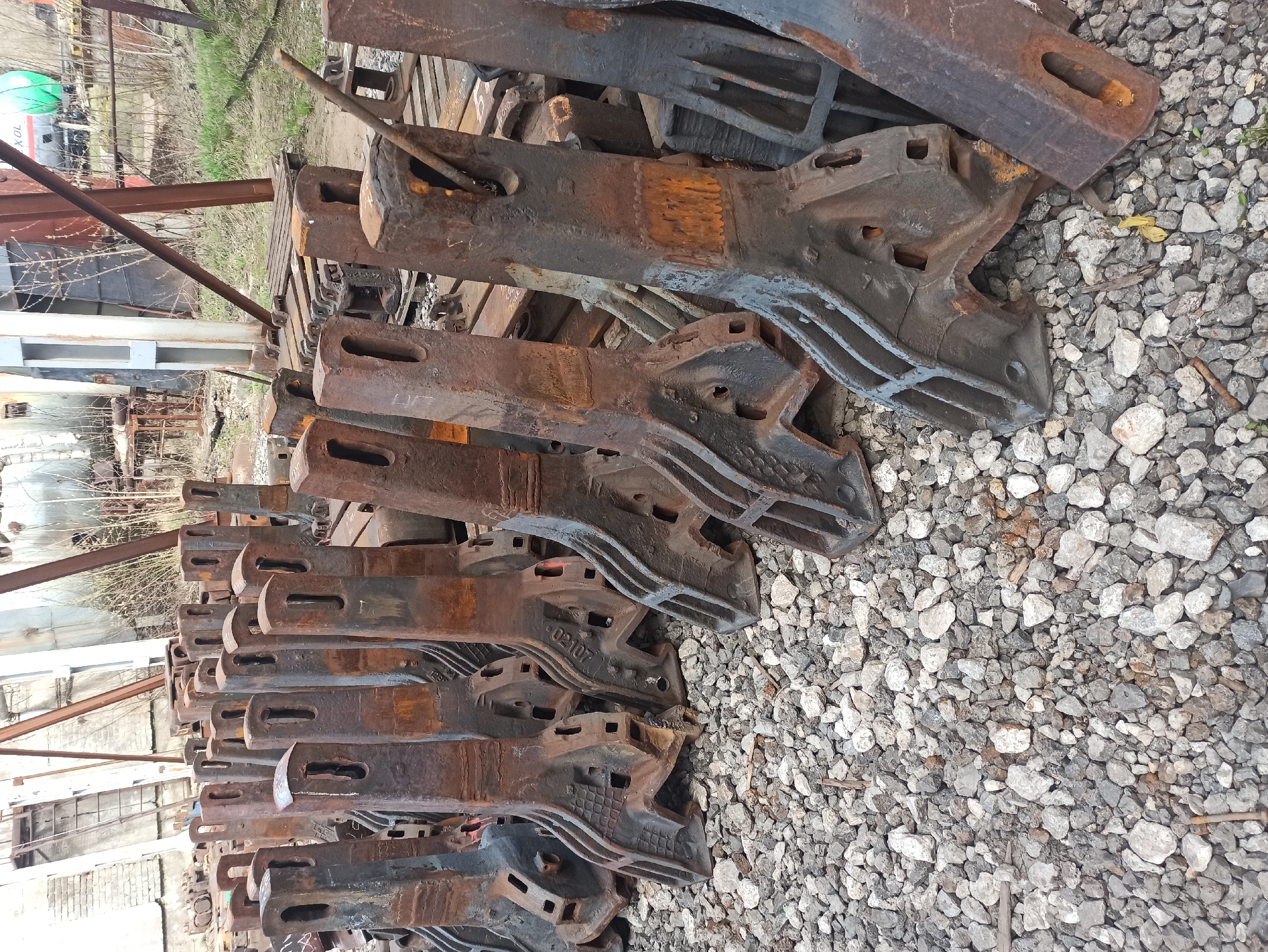The image shows a large pile of heavily rusted, possibly cast iron, railway or industrial machine parts. They are stacked on top of each other, some lying on a pallet, with a gravelly ground surface visible on the right side. The parts are likely from a railway car or similar heavy equipment, possibly for brakes or suspension, based on the shapes and slots. The setting is an industrial or storage area. While the location in Perm, Russia, cannot be conclusively confirmed from this image, the objects and environment are consistent with such a location. The pile appears abandoned and neglected. No people or actions are visible in the image.