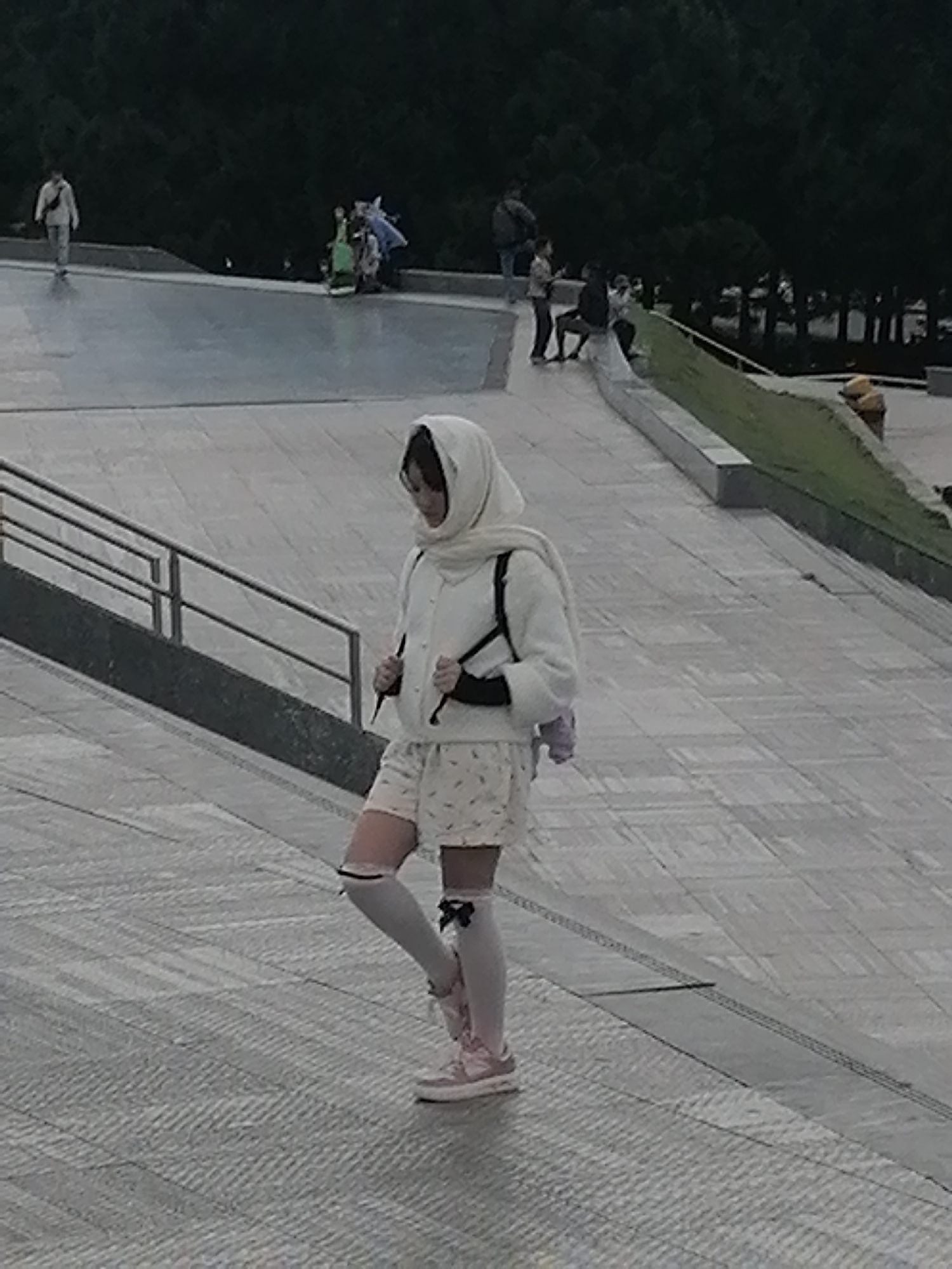 A young person, dressed in a white hooded jacket, patterned shorts, and white knee-high socks adorned with black bows, walks on a tiled pathway. They are carrying a dark bag with straps over their shoulders. The person is wearing pink sneakers. In the background, several other individuals are visible at a distance, some walking and others gathered in groups. The environment appears to be an outdoor public space with paved walkways, grassy areas, and trees. This location resembles the expansive outdoor areas of the Da Lat Flower Garden in Da Lat, Vietnam, known for its terraced landscapes and walking paths.