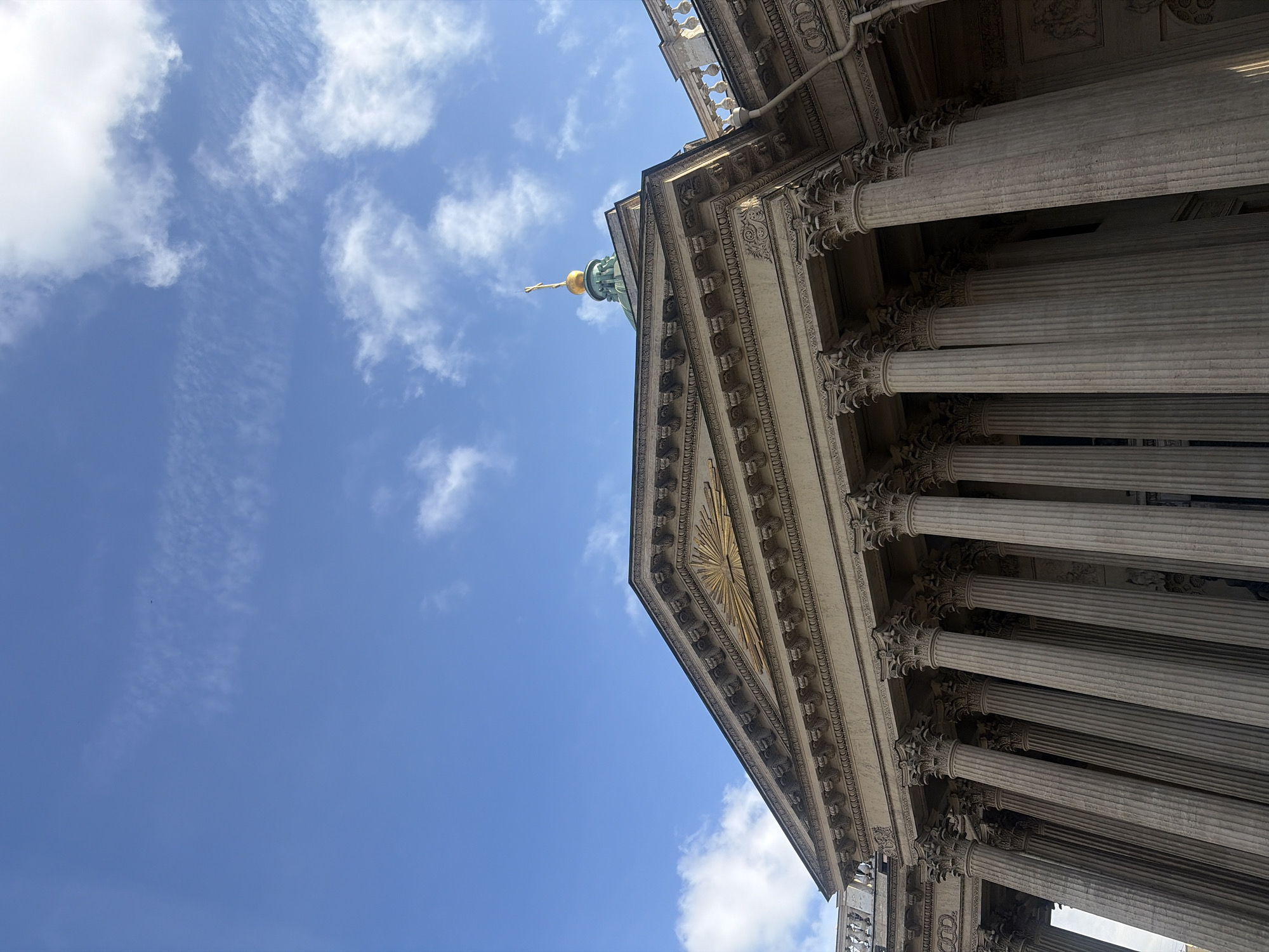 The image captures a low-angle view of the Kazan Cathedral in Saint Petersburg, Russia. The architecture features a grand colonnade of tall, fluted Corinthian columns supporting an ornate entablature and pediment. A golden sunburst motif is visible within the pediment. Above the main structure, a green dome topped with a golden cross is partially visible, hinting at the cathedral's religious significance. The sky is a clear blue, with scattered white clouds, suggesting a bright, possibly daytime, setting. The lighting casts shadows that emphasize the architectural details and the scale of the building. No people are visible in the frame. The overall impression is one of grandeur and historical significance, characteristic of St. Petersburg's imperial architecture.