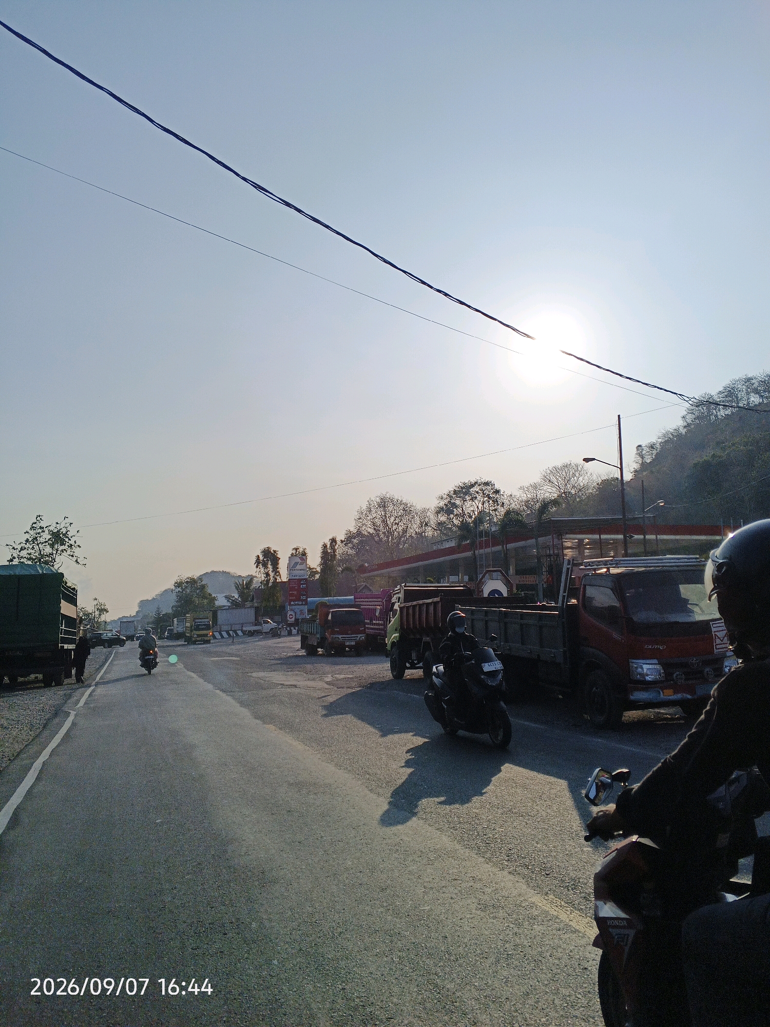 The image depicts a road scene in Pare-Pare, Indonesia, on September 7, 2026, at 16:44. The sun is low in the sky, casting long shadows and creating a bright glare. A traffic intersection appears to be the focus, with several vehicles present. In the foreground, two motorcycles are visible, one with a rider wearing a helmet and dark clothing, and another partially obscured. Several trucks, including a green flatbed truck and a red dump truck, are stopped or moving slowly. A gas station with a red awning and a sign for "Pertamina" is visible on the right side of the road. Trees and a hilly landscape form the background, with power lines stretching across the sky. The road appears to be asphalt with white lane markings.