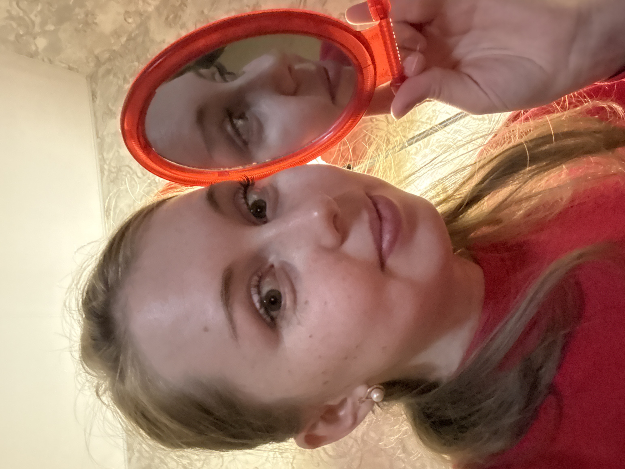 A young woman is depicted in a close-up, upward-angled shot, looking directly at the camera with a calm expression. She holds a small, red, oval-framed mirror with a handle above her head, reflecting her own face back towards the viewer. Her reflection in the mirror also shows her gazing in the same direction.
The woman has long, light blonde hair that cascades around her shoulders and is illuminated by a warm light source, creating bright highlights. She has fair skin, with visible freckles across her nose and cheeks. Her eyes are light brown, and she wears a single pearl earring in her visible right ear. She is dressed in a red garment, possibly a sweater or top, which matches the color of the mirror frame.
The background suggests an indoor setting, with a light-colored wall featuring a subtle, possibly floral or textured, pattern. The lighting appears to be artificial and warm, casting a gentle glow on the scene. There is no visible text in the image.