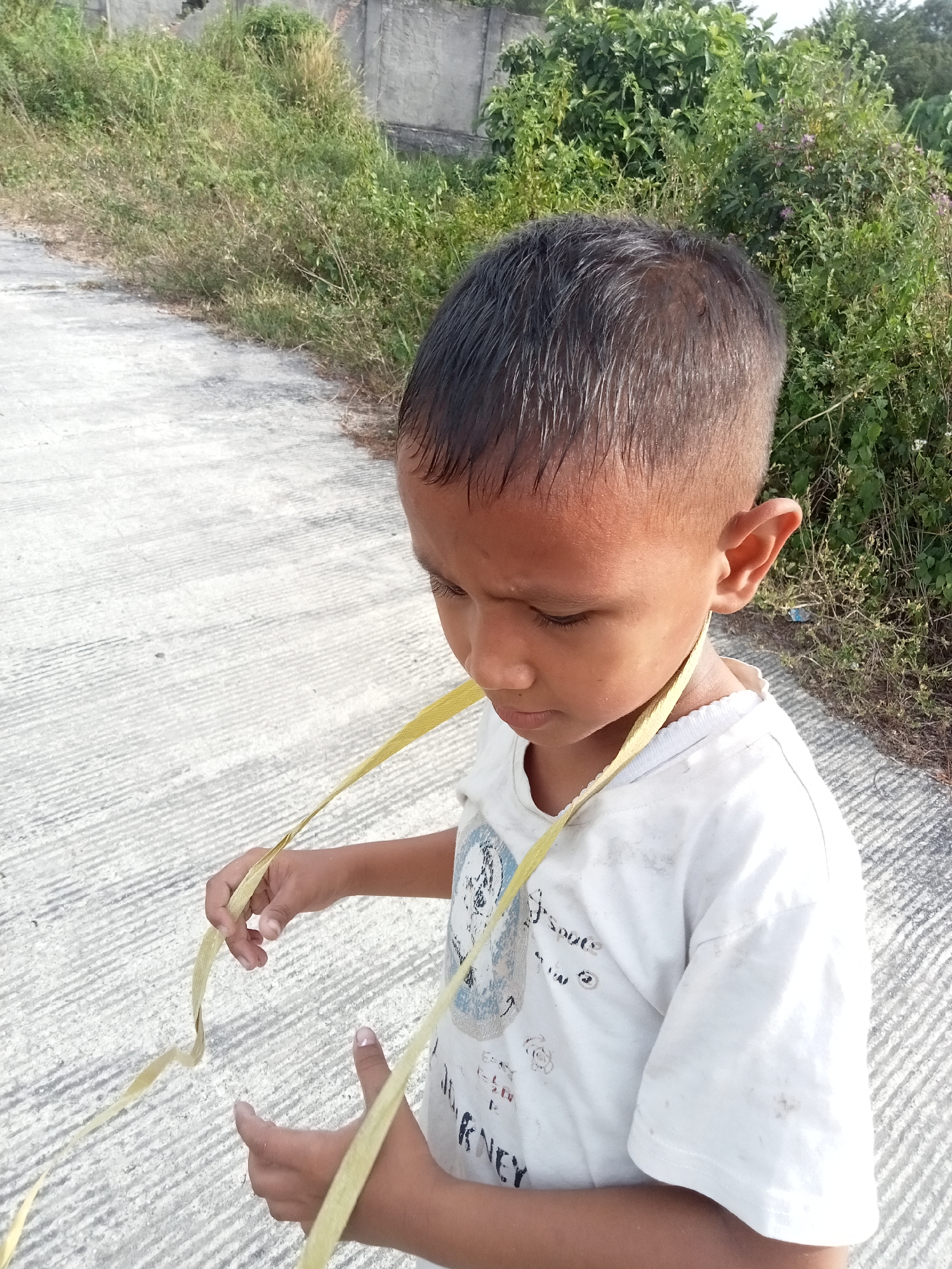 A young boy with dark hair and brown eyes is shown from the chest up, looking down and to his right. He is wearing a white t-shirt with a graphic design and a yellow ribbon or strap is around his neck and draped over his shirt. The boy's hands are visible, holding the yellow object. He is standing on a concrete path that has a ridged texture. To the right of the path is a dense area of green vegetation and foliage, with some dry grass and a grey wall in the background. The scene appears to be outdoors, with natural lighting. This photograph was taken in Kualu, Indonesia.